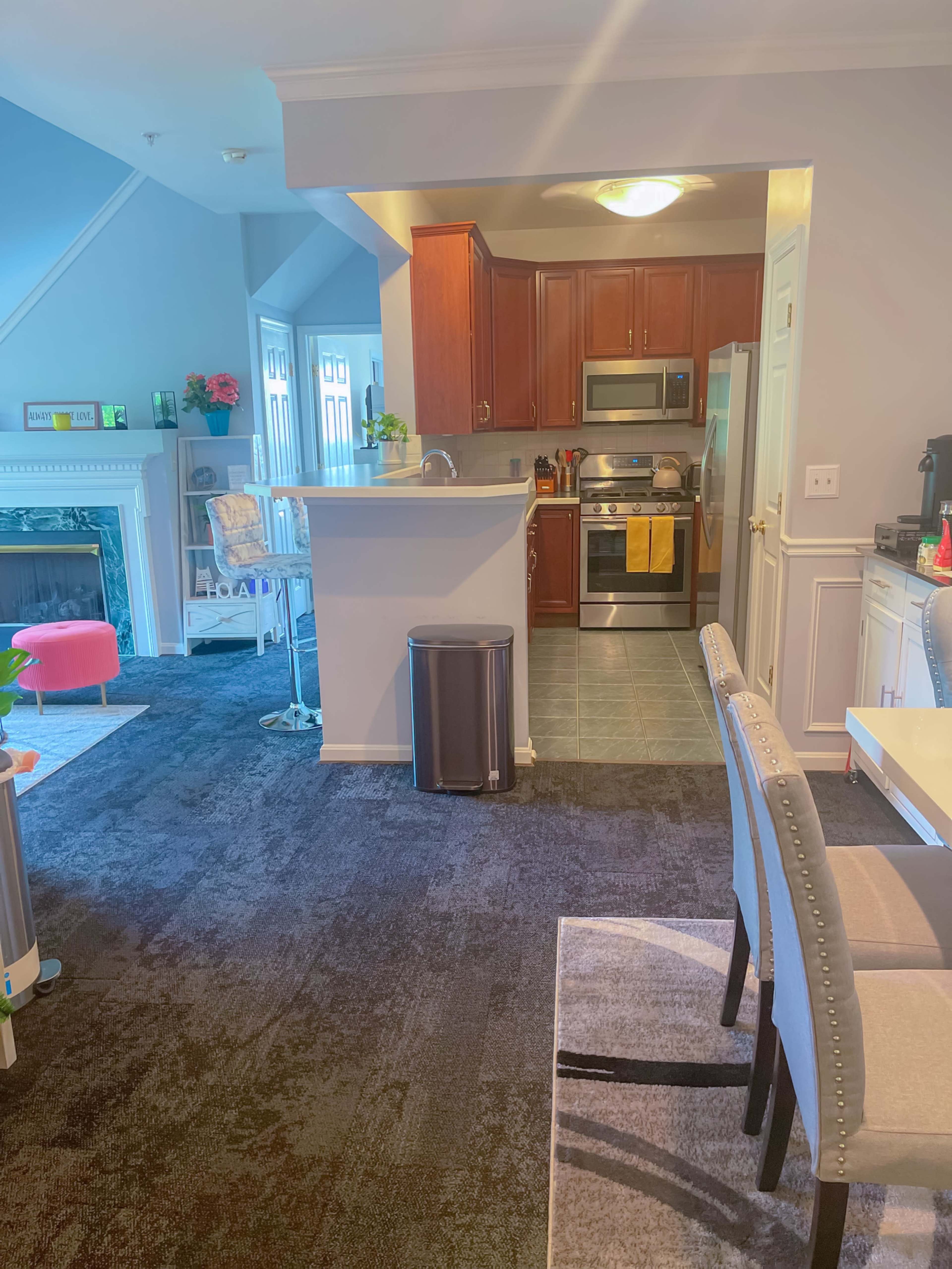 A modern kitchen area with wood cabinets, stainless steel appliances, and a dining space adjacent to it, featuring a table and chairs on a dark carpet.