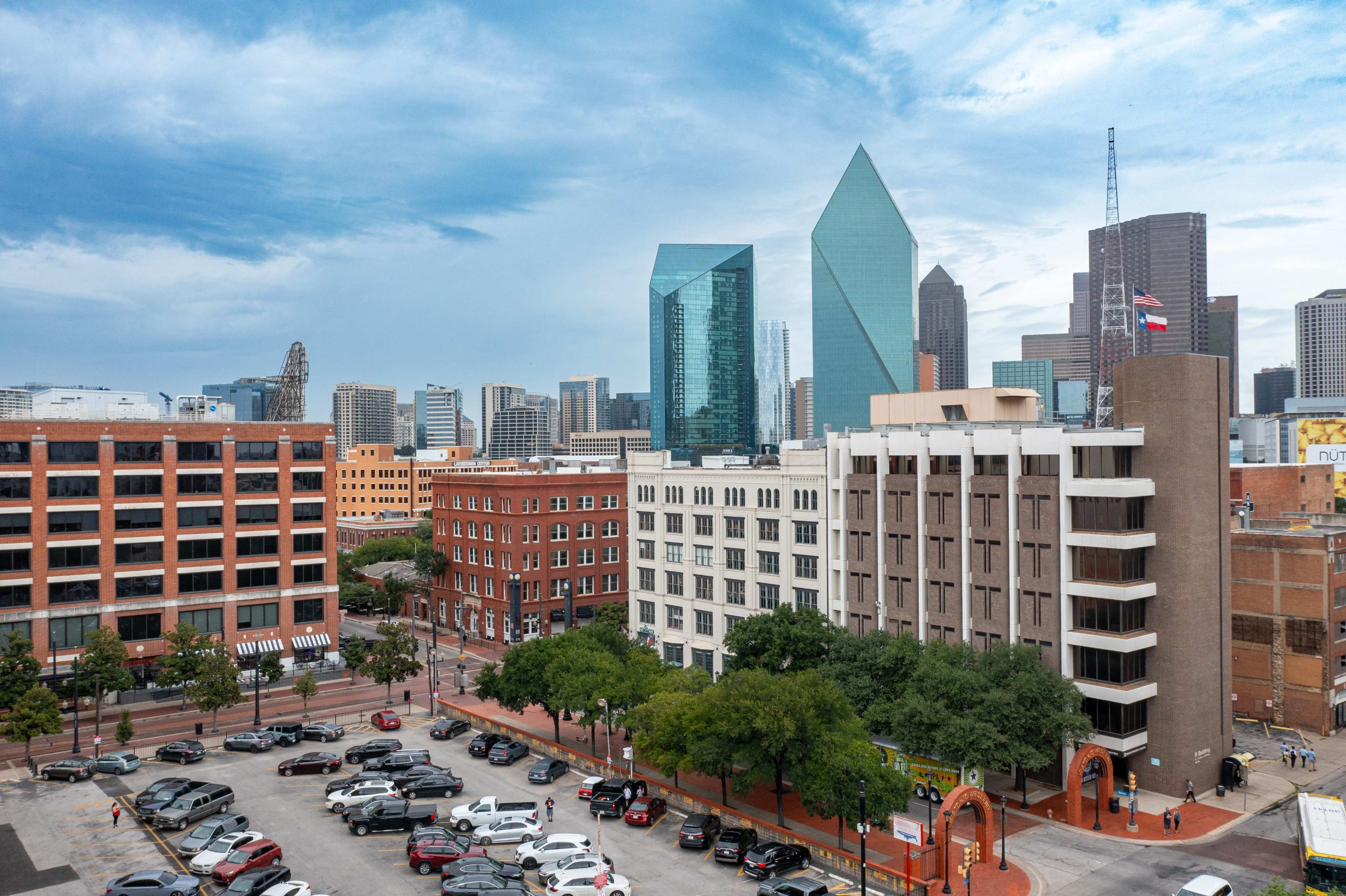 The image shows a view of a city skyline with modern glass buildings, including a distinctive diamond-shaped structure, alongside low-rise brick buildings and parked cars on the street below.