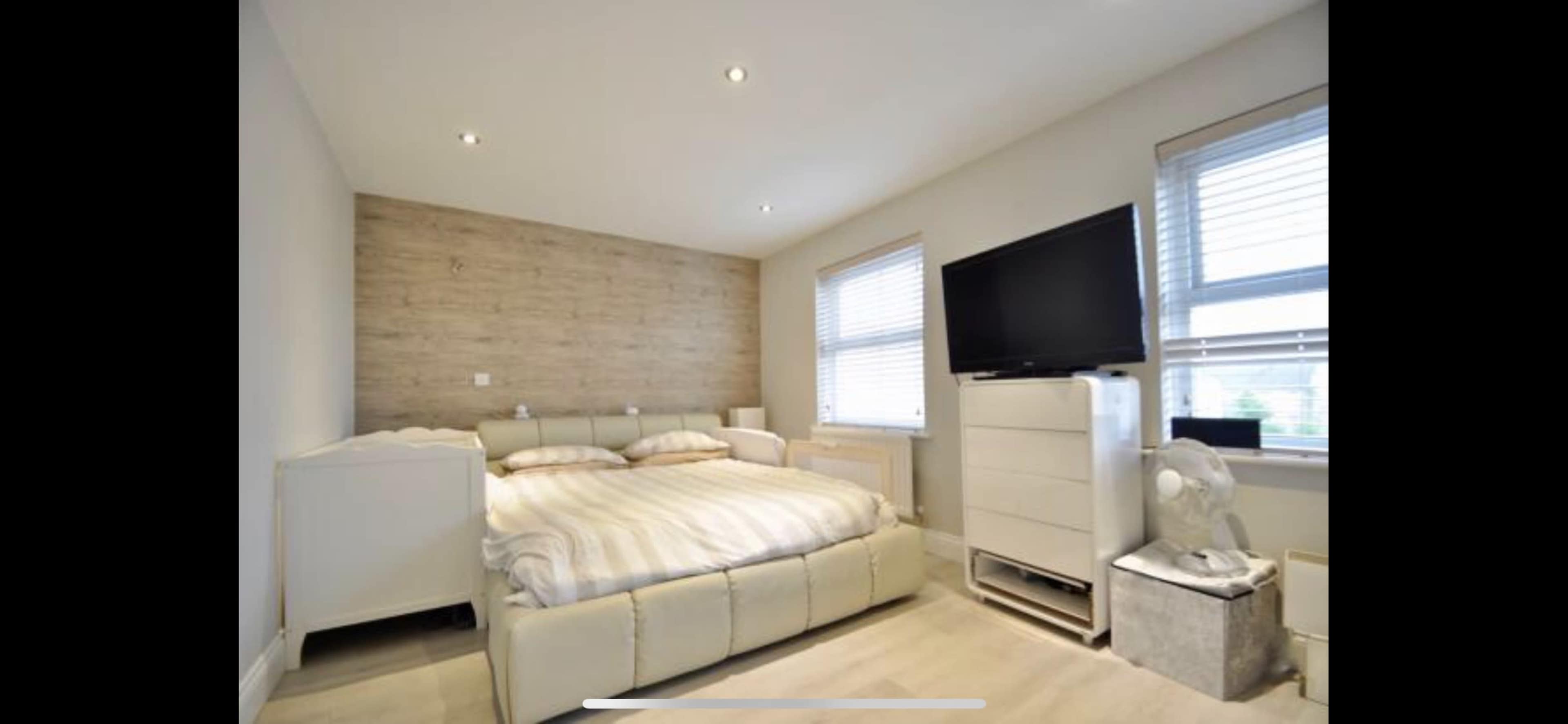 A bedroom with a large bed, a television mounted on the wall, and two windows allowing natural light in.
