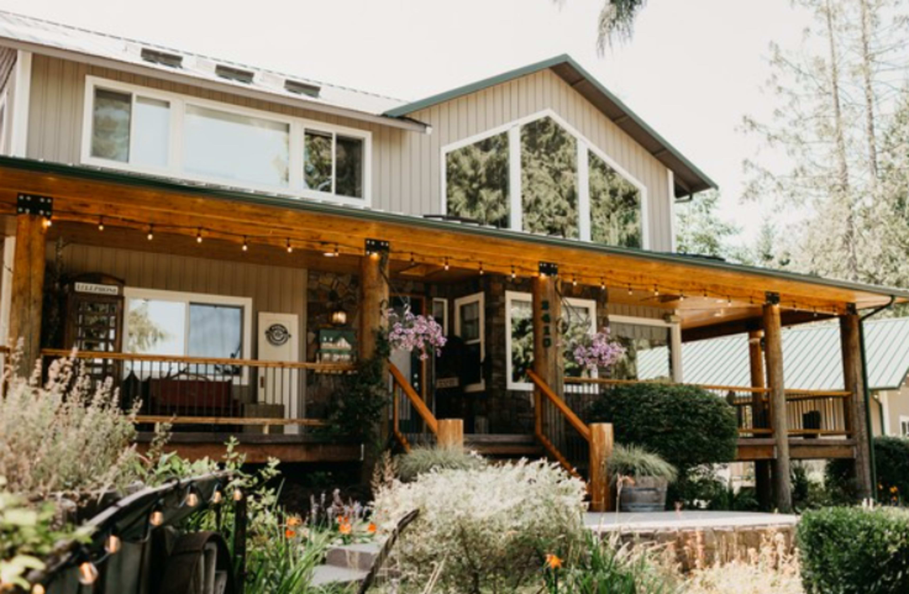 The image shows a two-story house with a wide wooden porch, large windows, and a landscaped front yard featuring flowers and greenery.