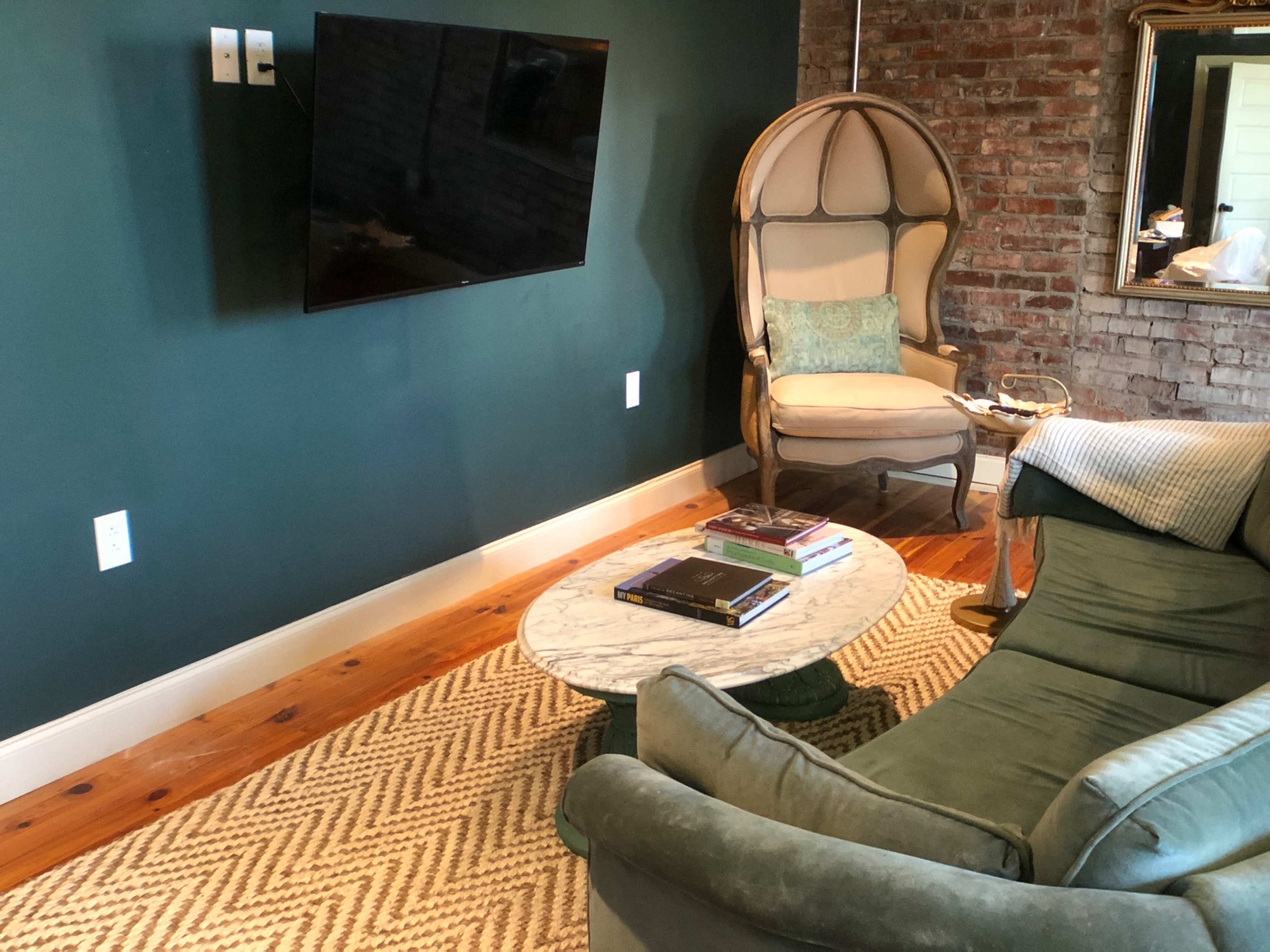 A cozy living space features a green wall, a mounted TV, a vintage chair, and a marble coffee table surrounded by a sofa and a woven rug.