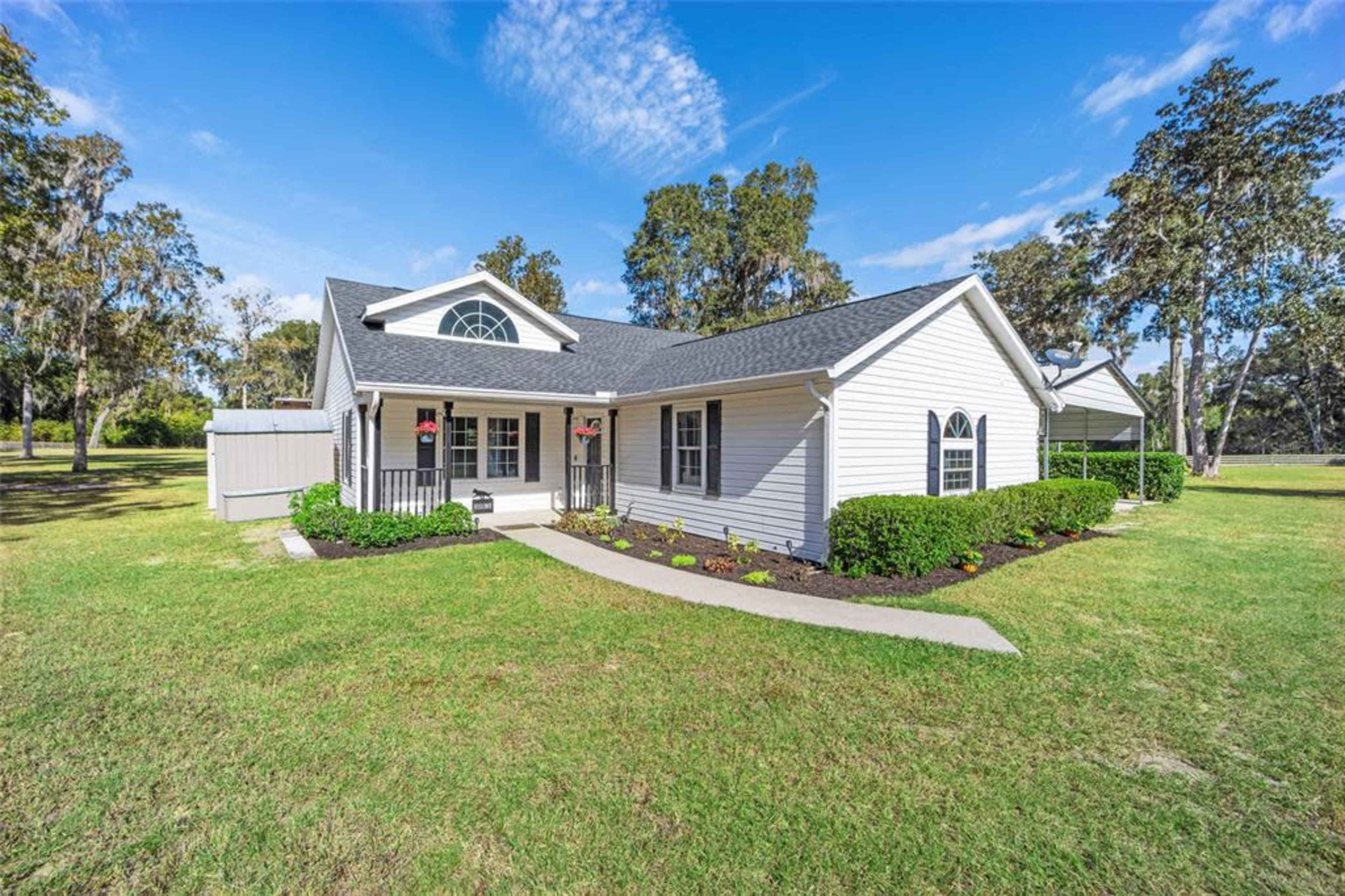 The image shows a single-story, light-colored house with a gabled roof, surrounded by a well-maintained lawn and landscaped bushes.