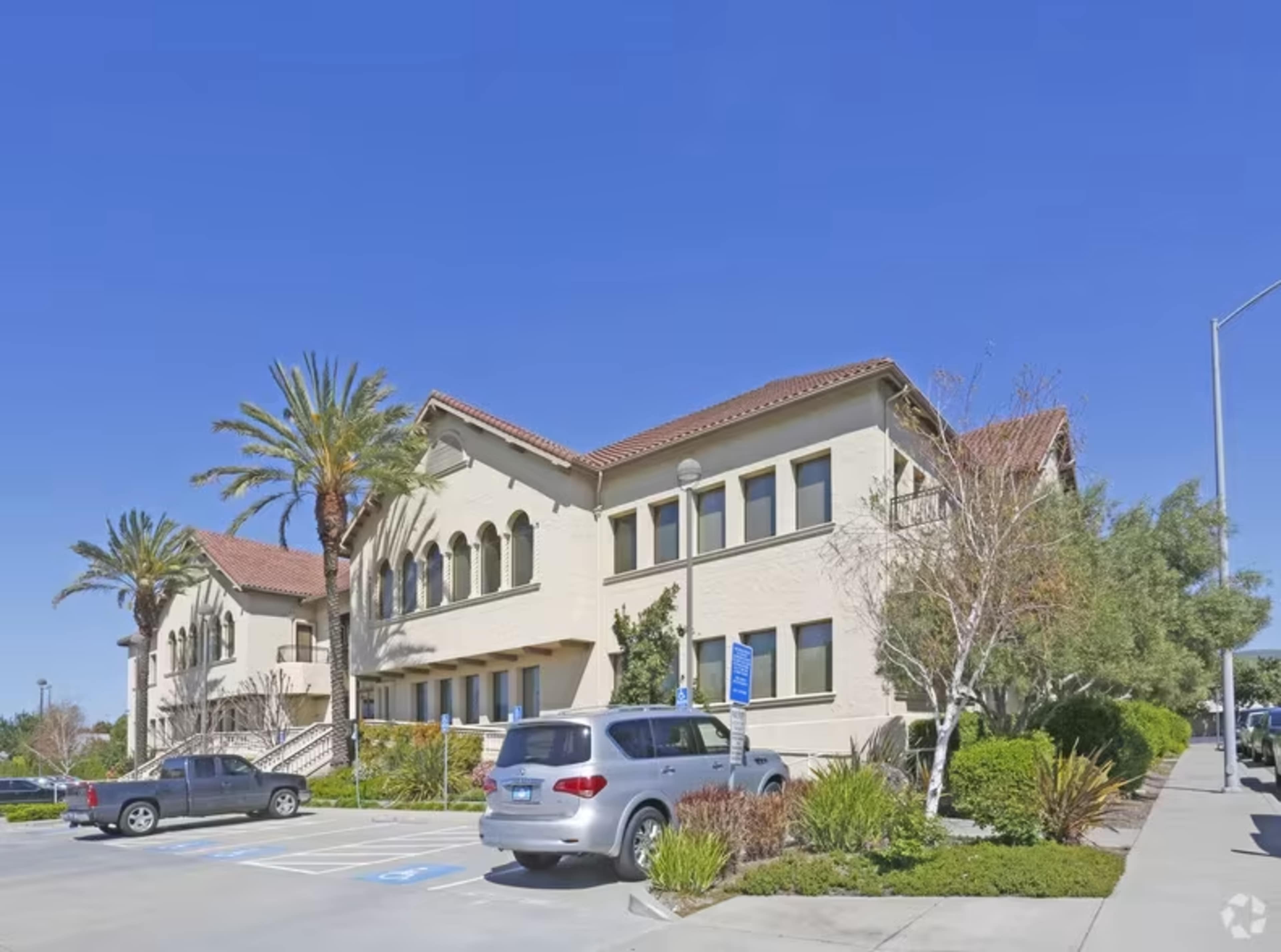 A two-story building with a tile roof is surrounded by palm trees and shrubs in a well-maintained parking lot.