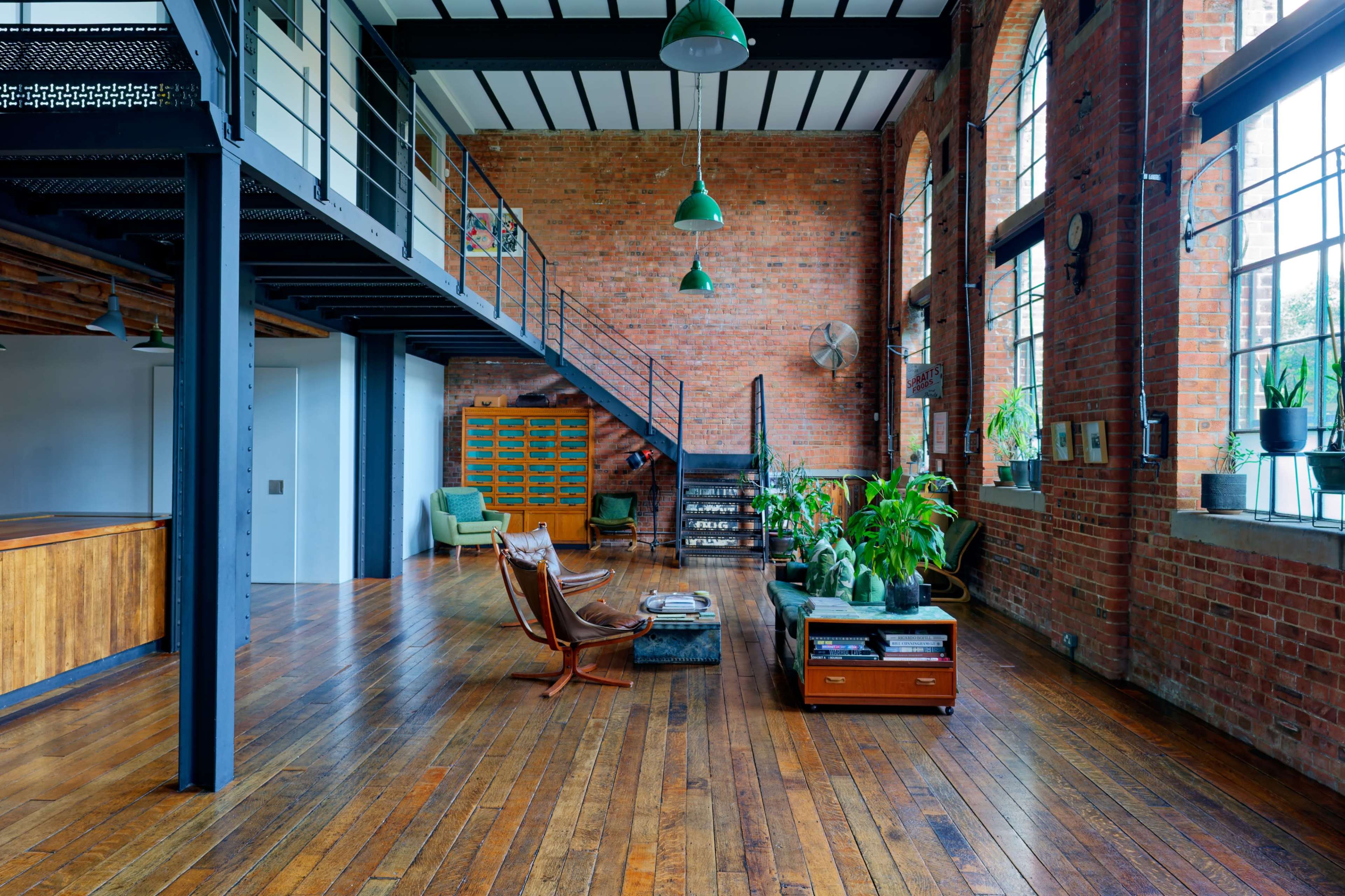 The image shows a spacious industrial-style loft featuring exposed brick walls, large windows, wooden floors, and a mix of modern and vintage furniture.