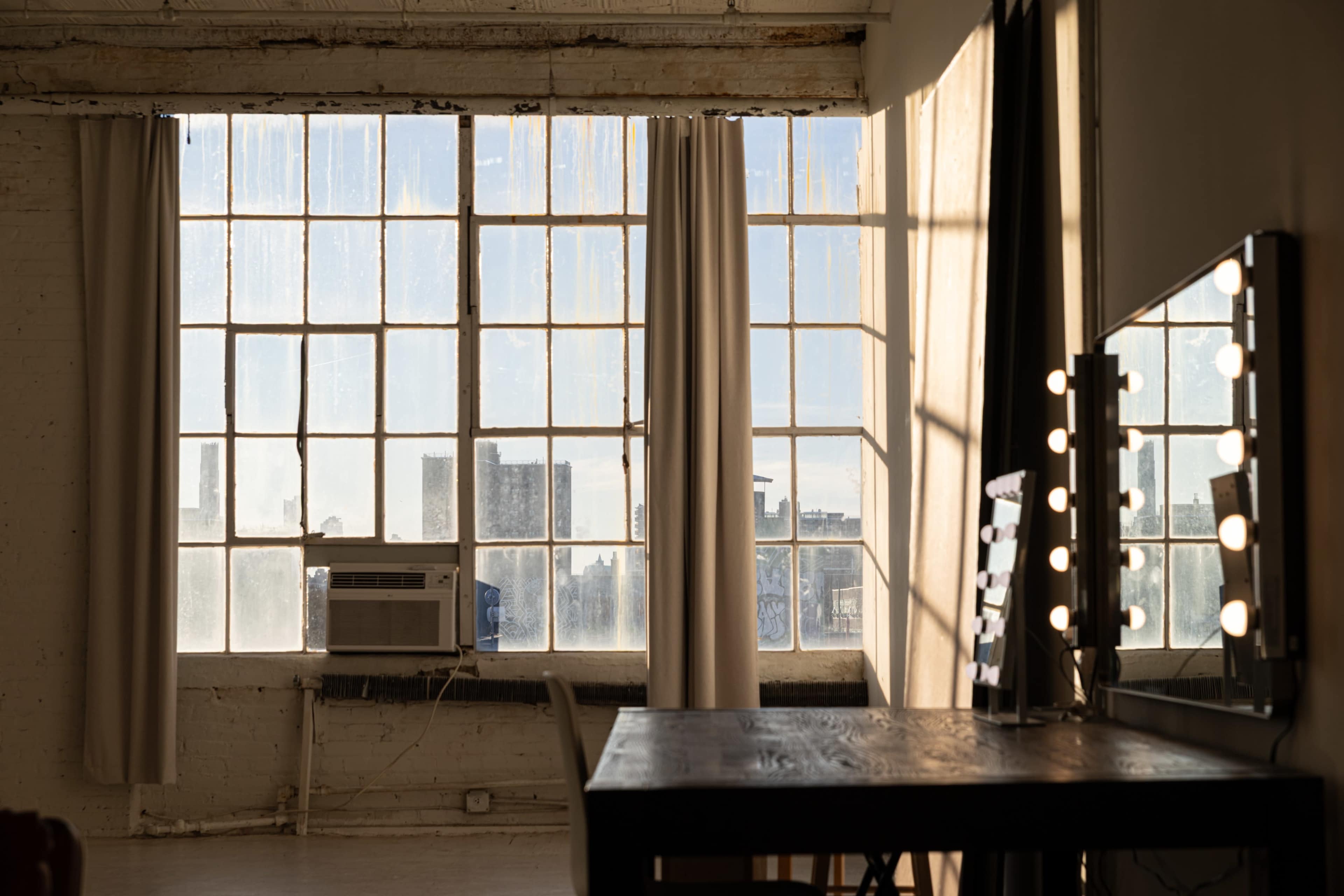 The image shows a sunlit room with large, dirty windows, a table, and a mirror with lights against one wall.