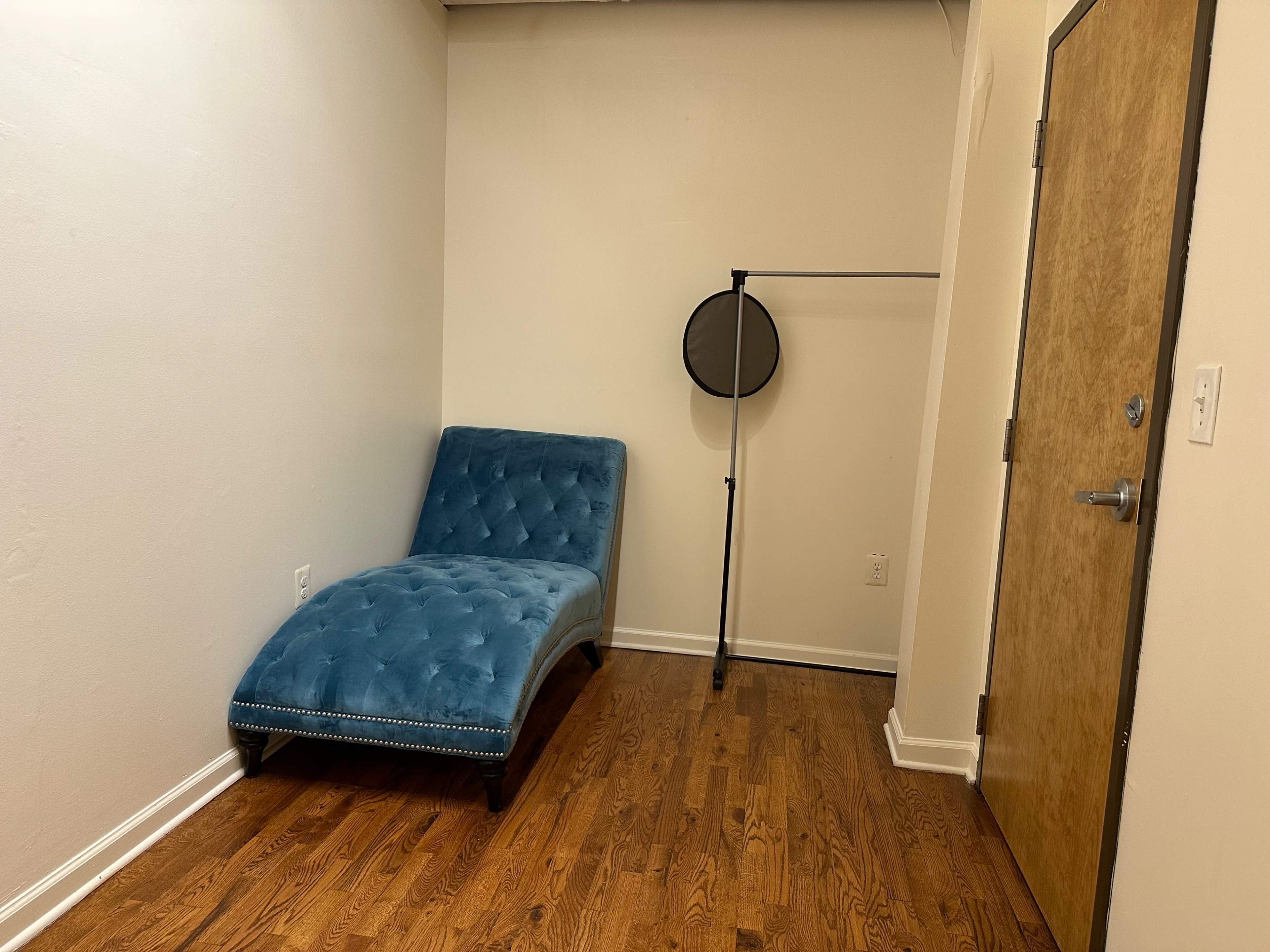 The image shows a small room with a blue chaise lounge and a metal clothing rack against a plain wall.
