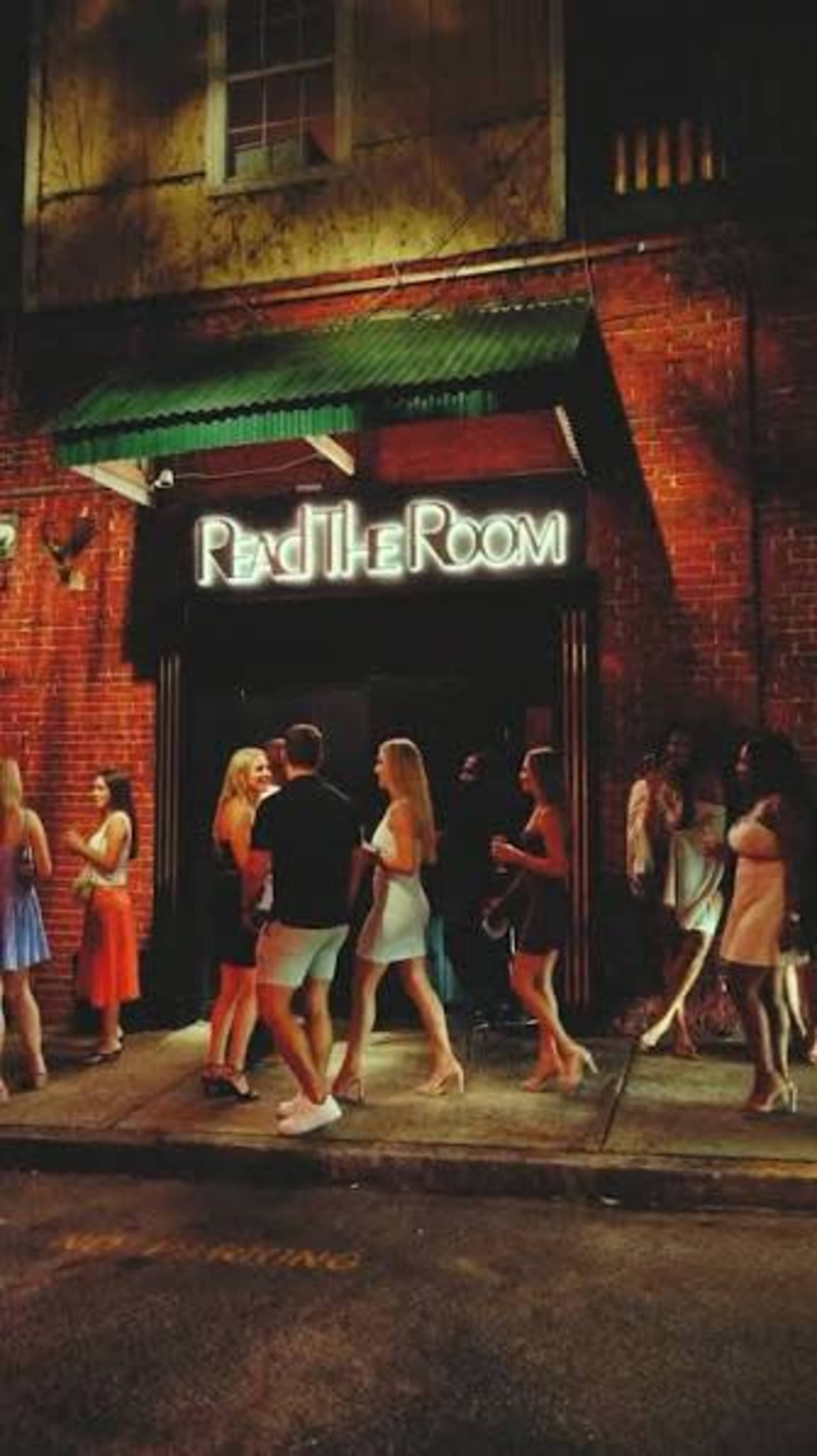 A group of people dressed in evening attire gathers outside a venue with a glowing sign that reads "Read The Room."