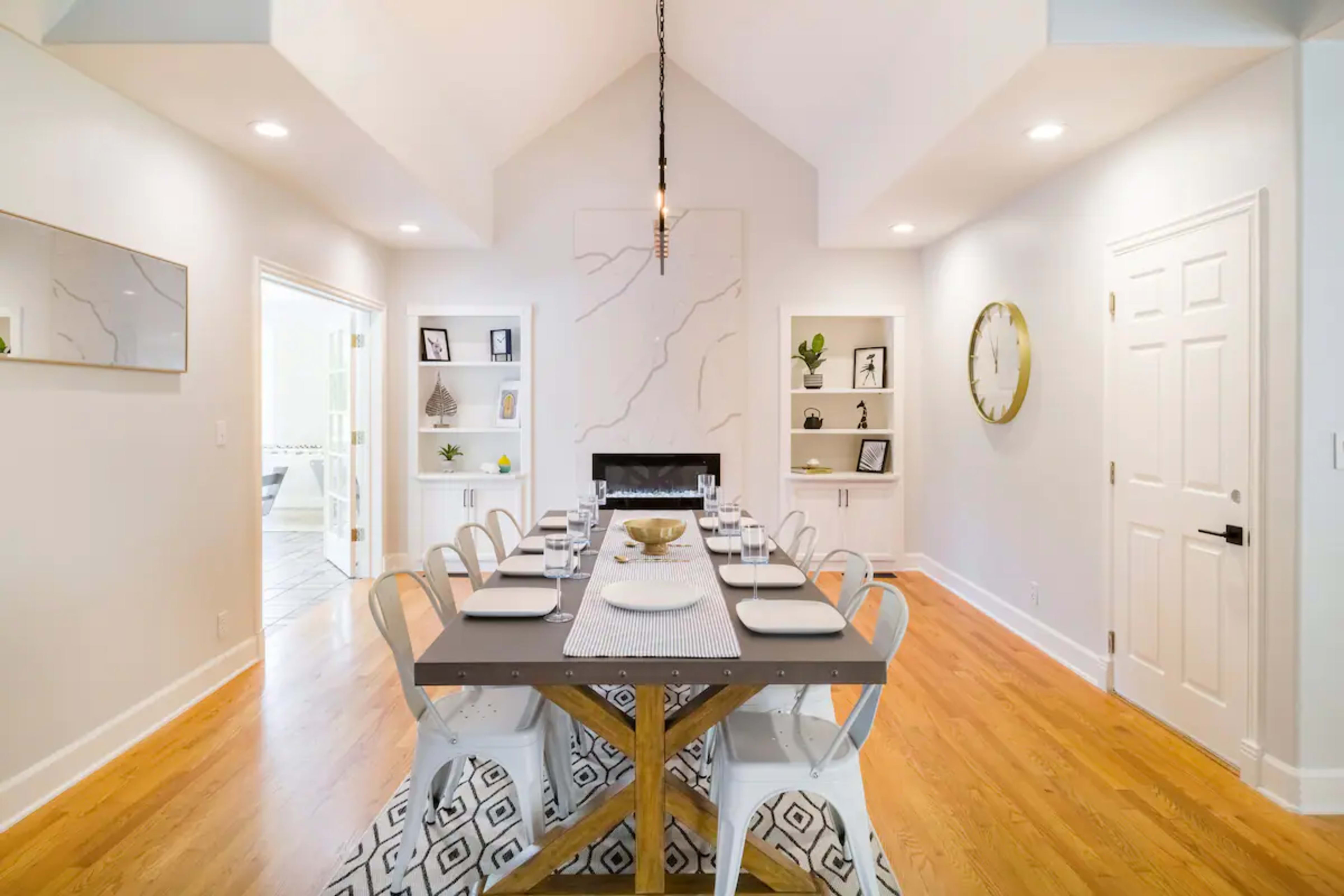 A modern dining room features a long table set with plates and utensils, surrounded by white chairs, with a decorative wall and a small fireplace in the background.
