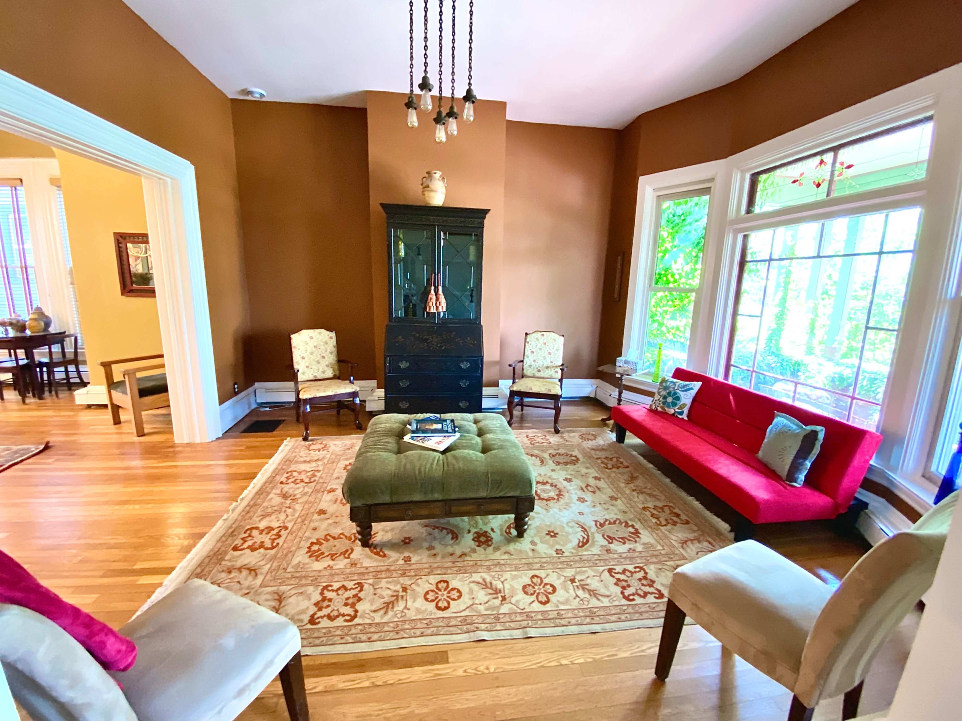 A living room with a red sofa, four chairs arranged around a green ottoman, and large windows allowing natural light to enter.