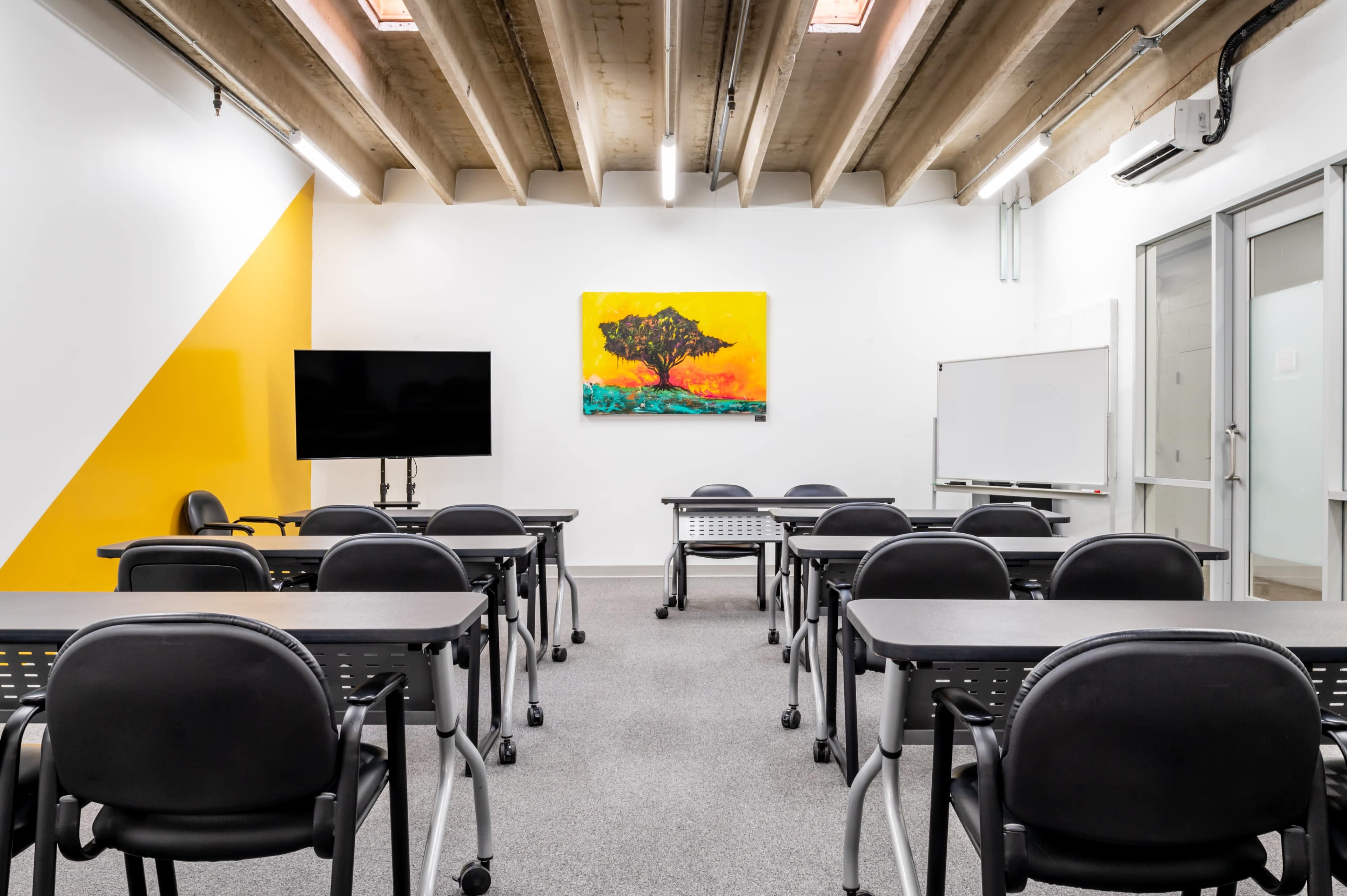 A classroom is arranged with black desks and chairs facing a large wall-mounted screen and a colorful artwork of a tree.