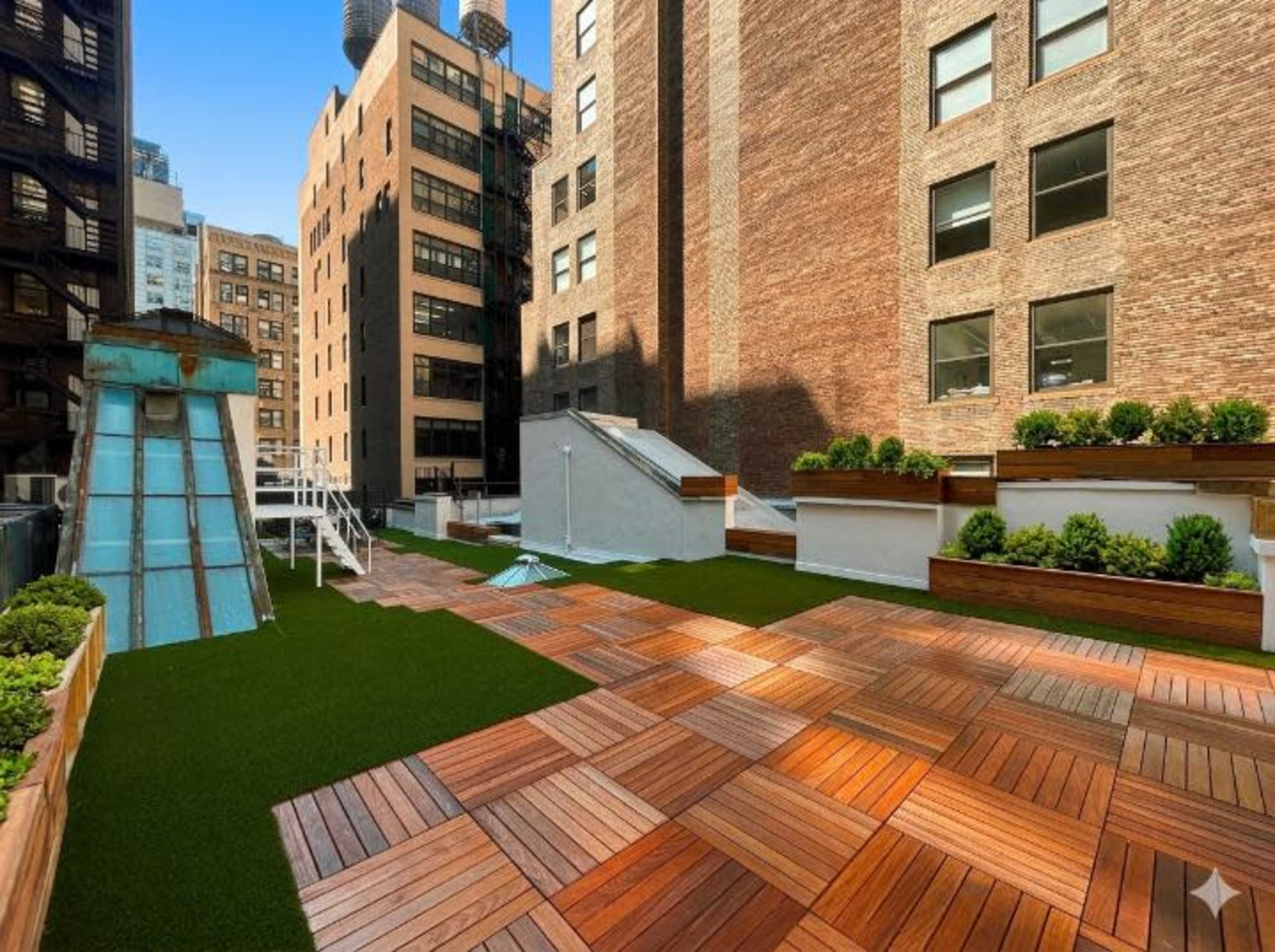 The image shows a rooftop area featuring wooden decking, lush greenery, and a small slide.
