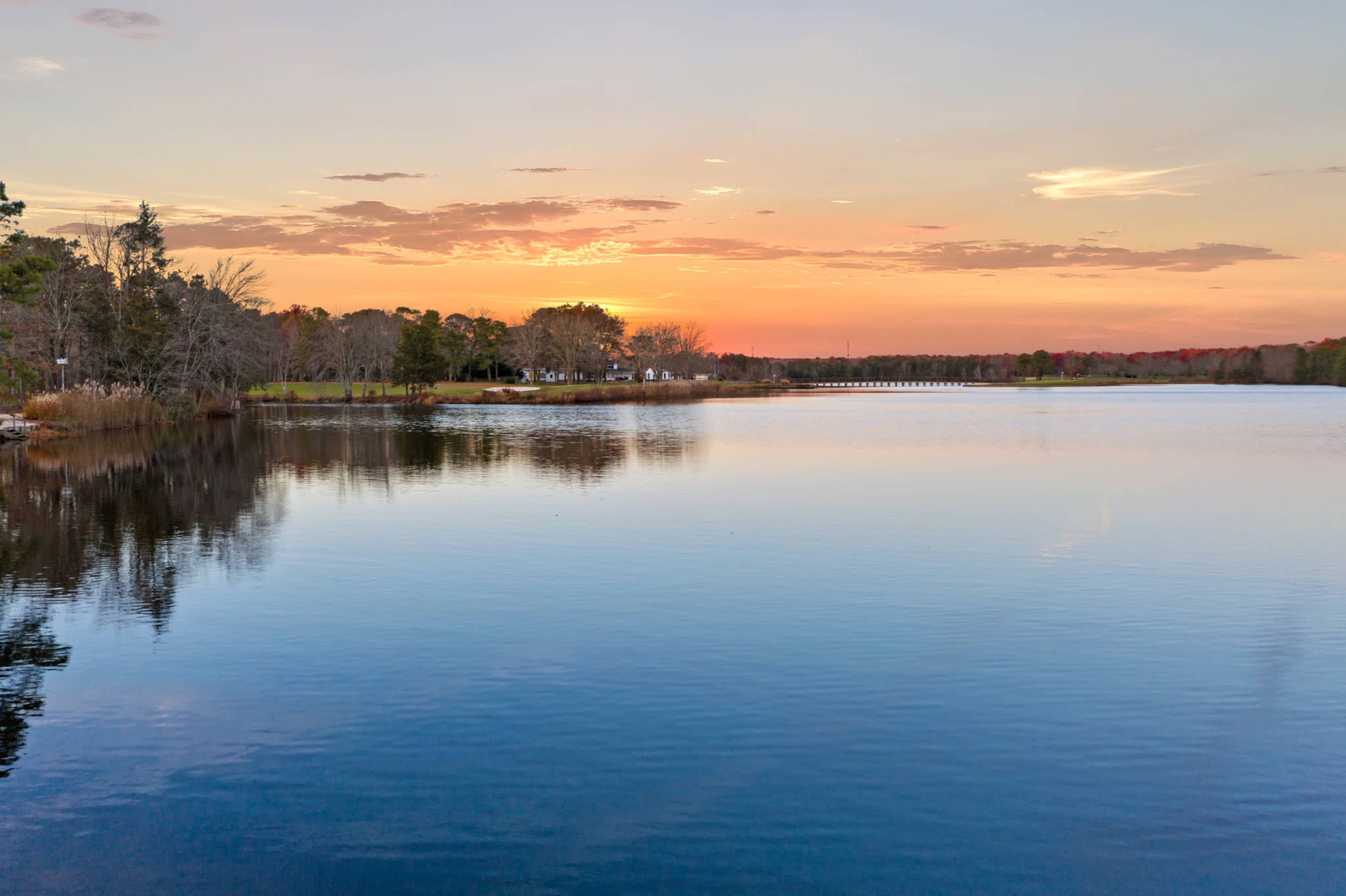 A calm lake reflecting a colorful sunset, surrounded by trees and distant buildings.