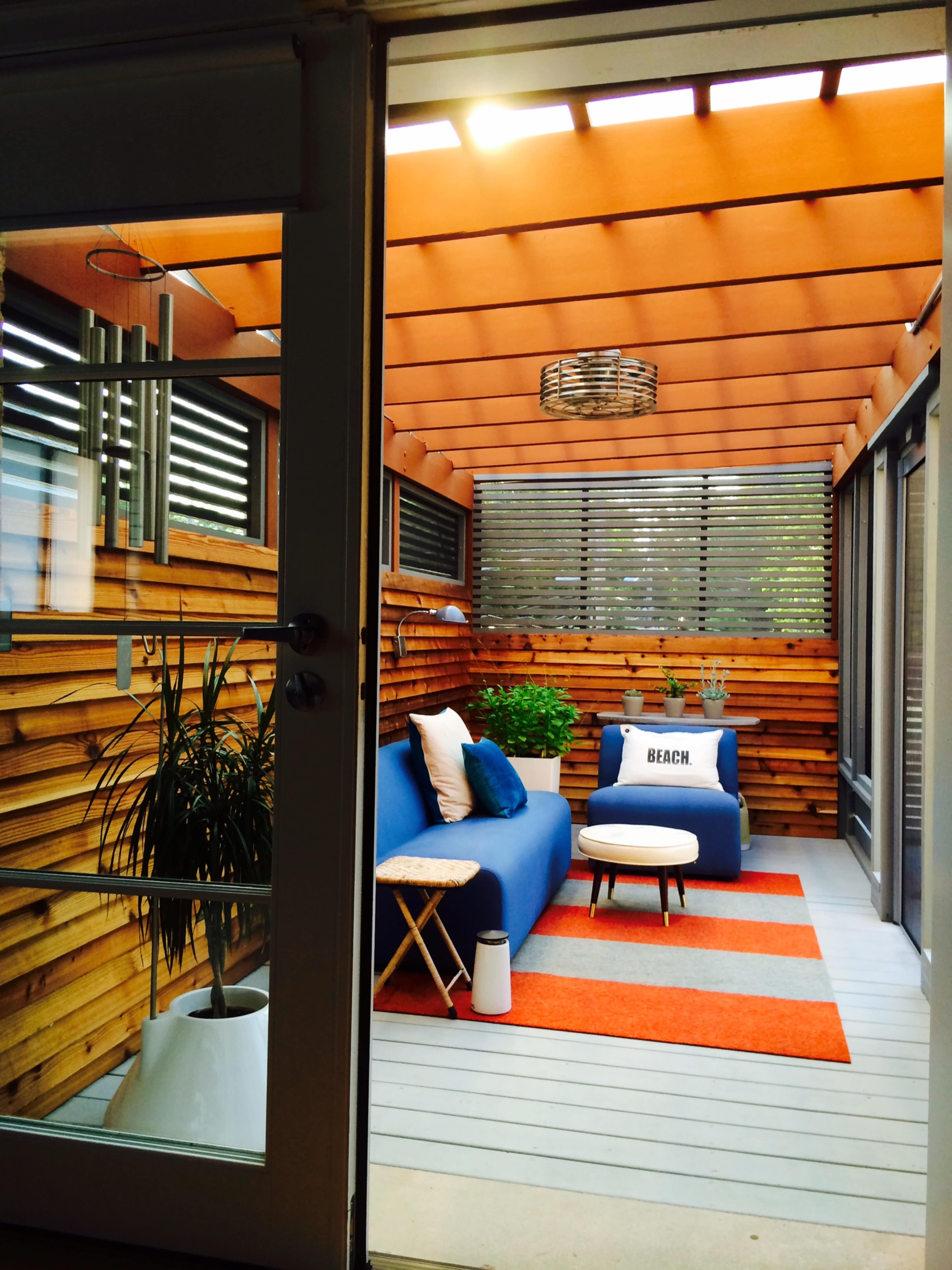 The image shows a cozy indoor seating area with a blue sofa, patterned rug, and wooden walls, illuminated by natural light coming through slatted windows.
