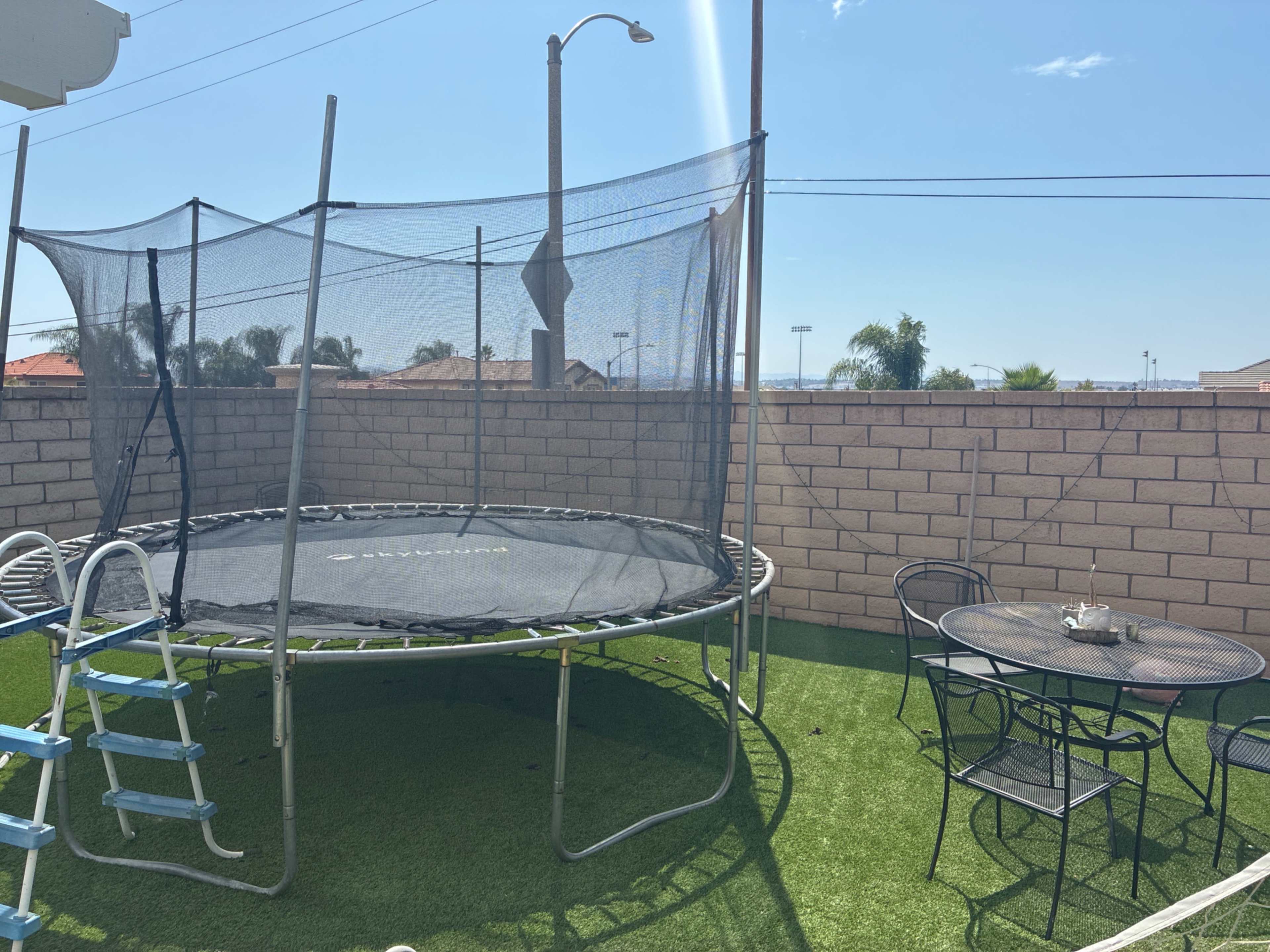 A trampoline with a net enclosure sits on green artificial grass next to a small round table and chairs in a backyard setting.