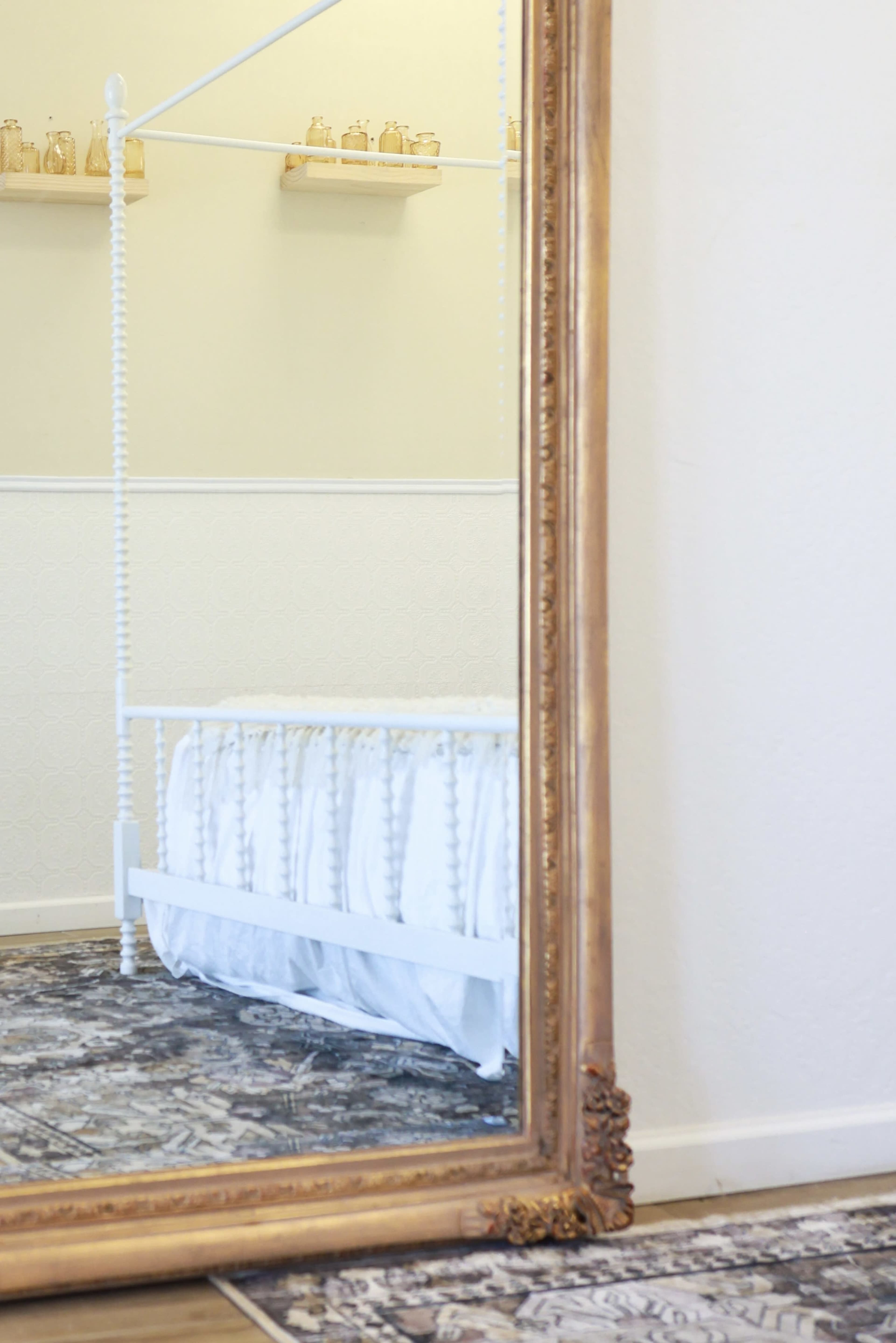 A large, ornate mirror reflects a white bed with a ruffled coverlet and a patterned rug on the floor.