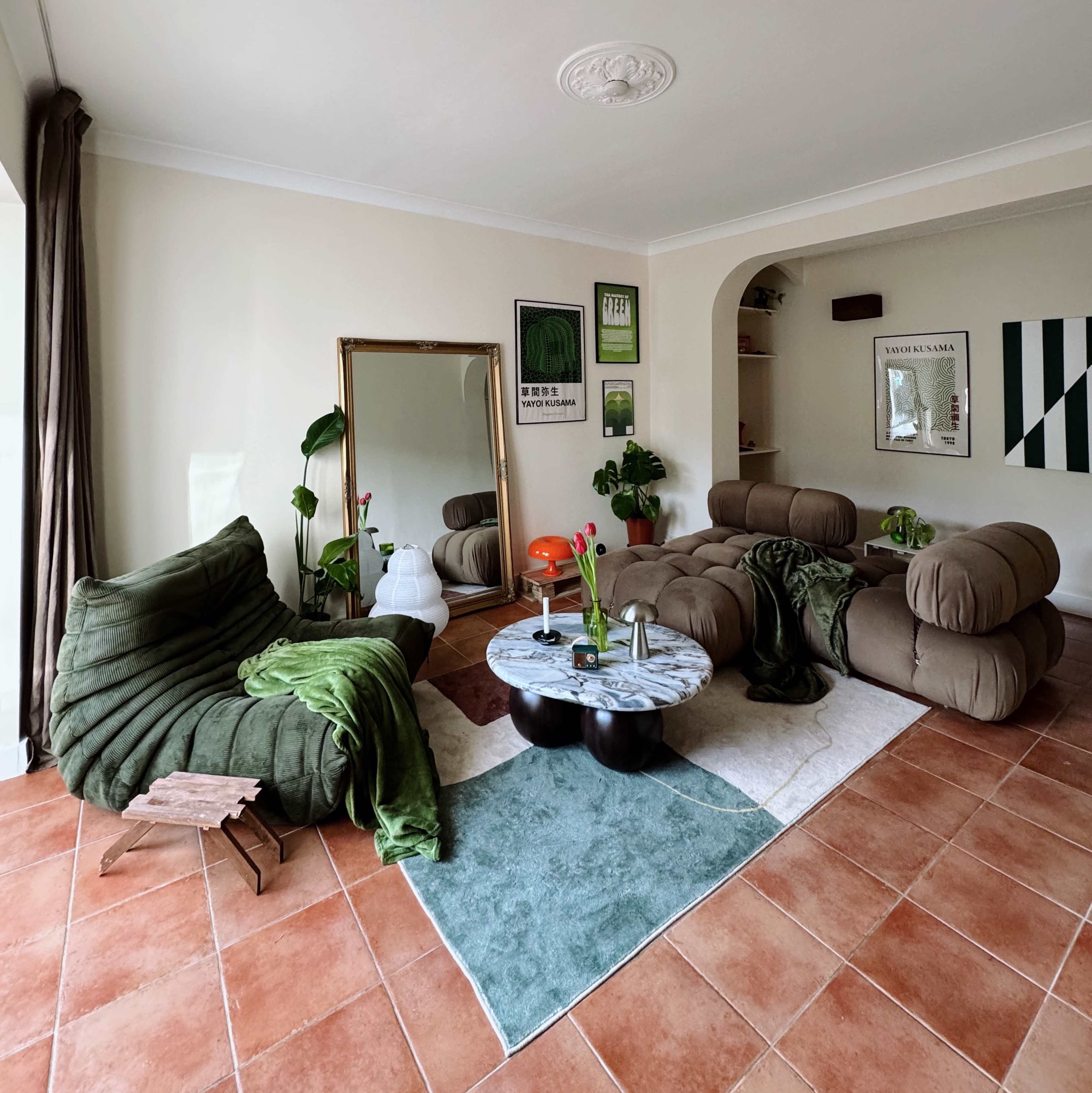 The living room features two modern sofas, a round marble coffee table, and plants, arranged on a patterned area rug with terracotta tile flooring.