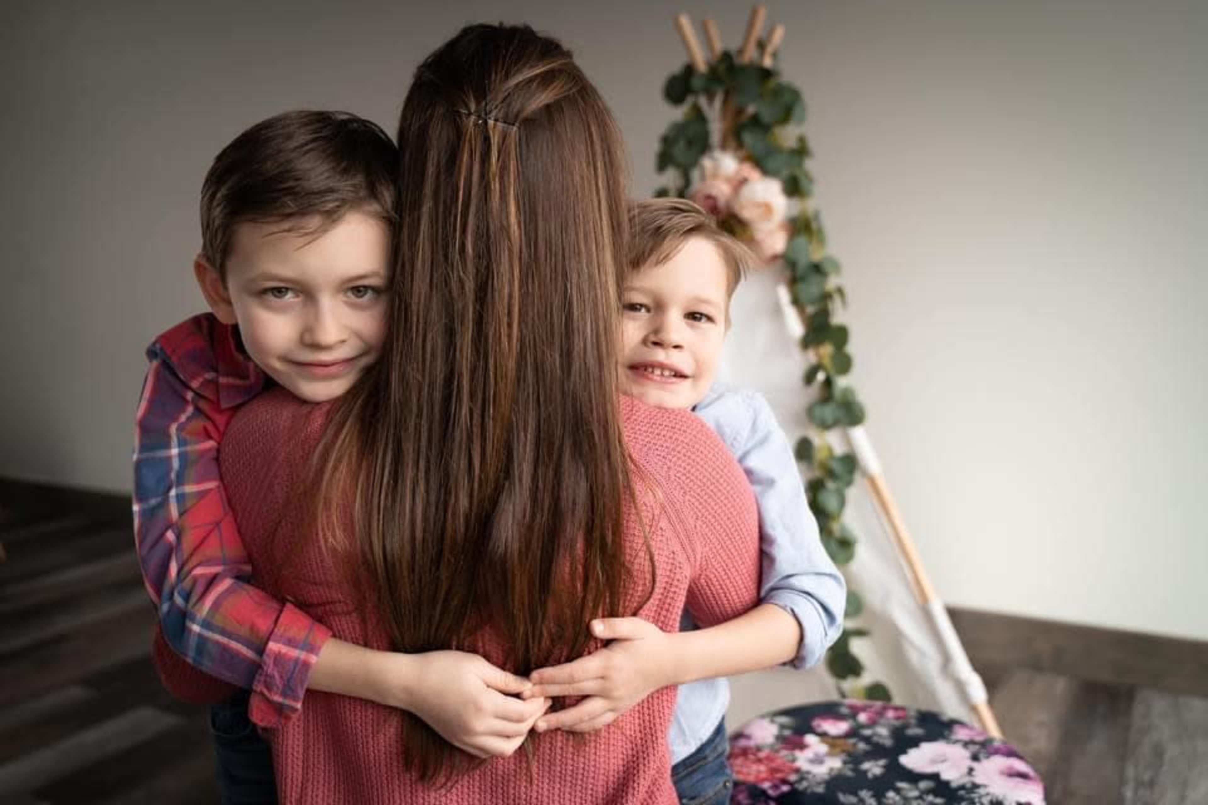 A woman stands with her two children, who are hugging her from behind, in a room decorated with flowers and greenery.