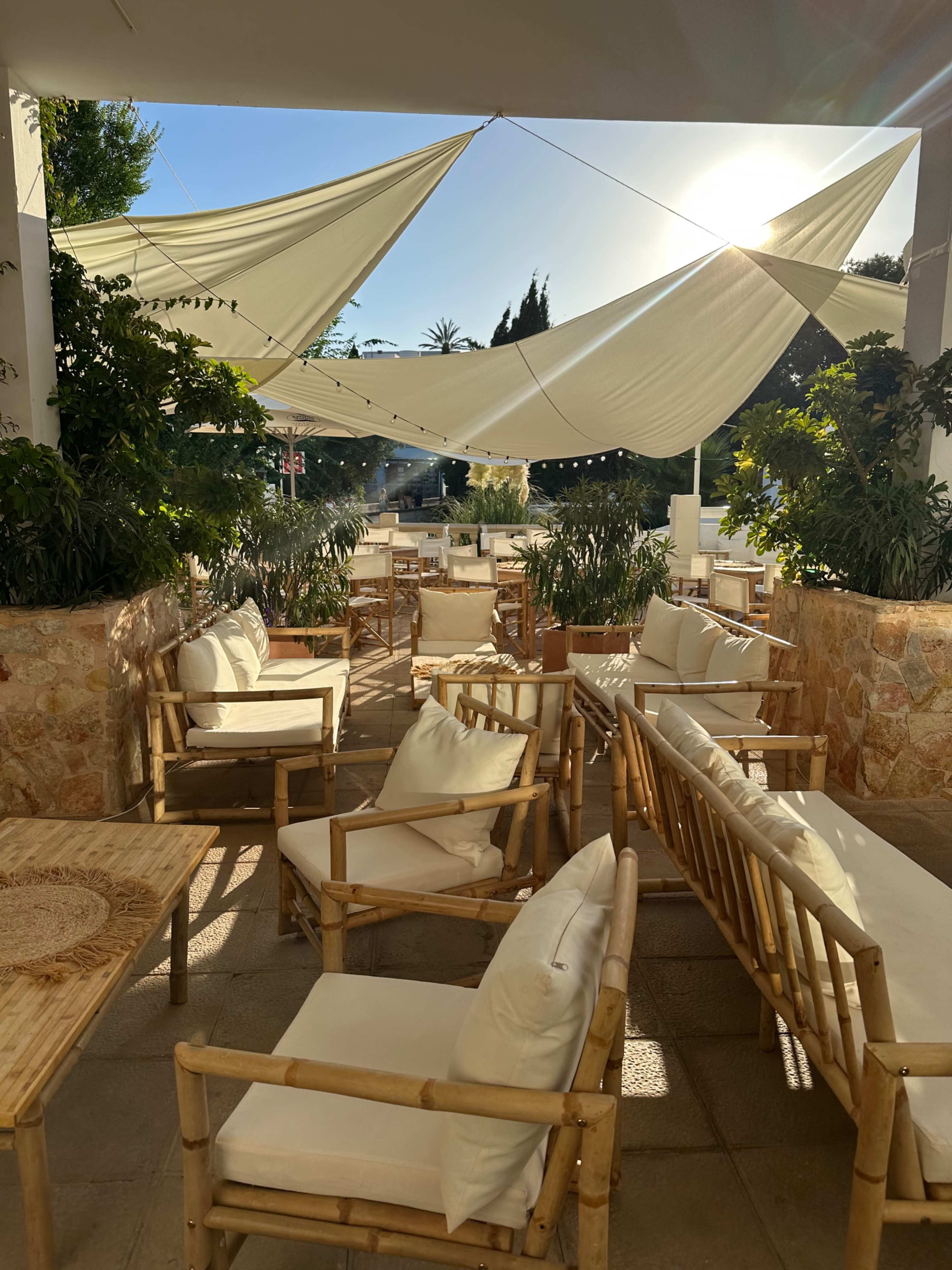 A bamboo-furnished outdoor seating area features white cushions and decorative plants under large, slanted canopies with sunlight streaming through.