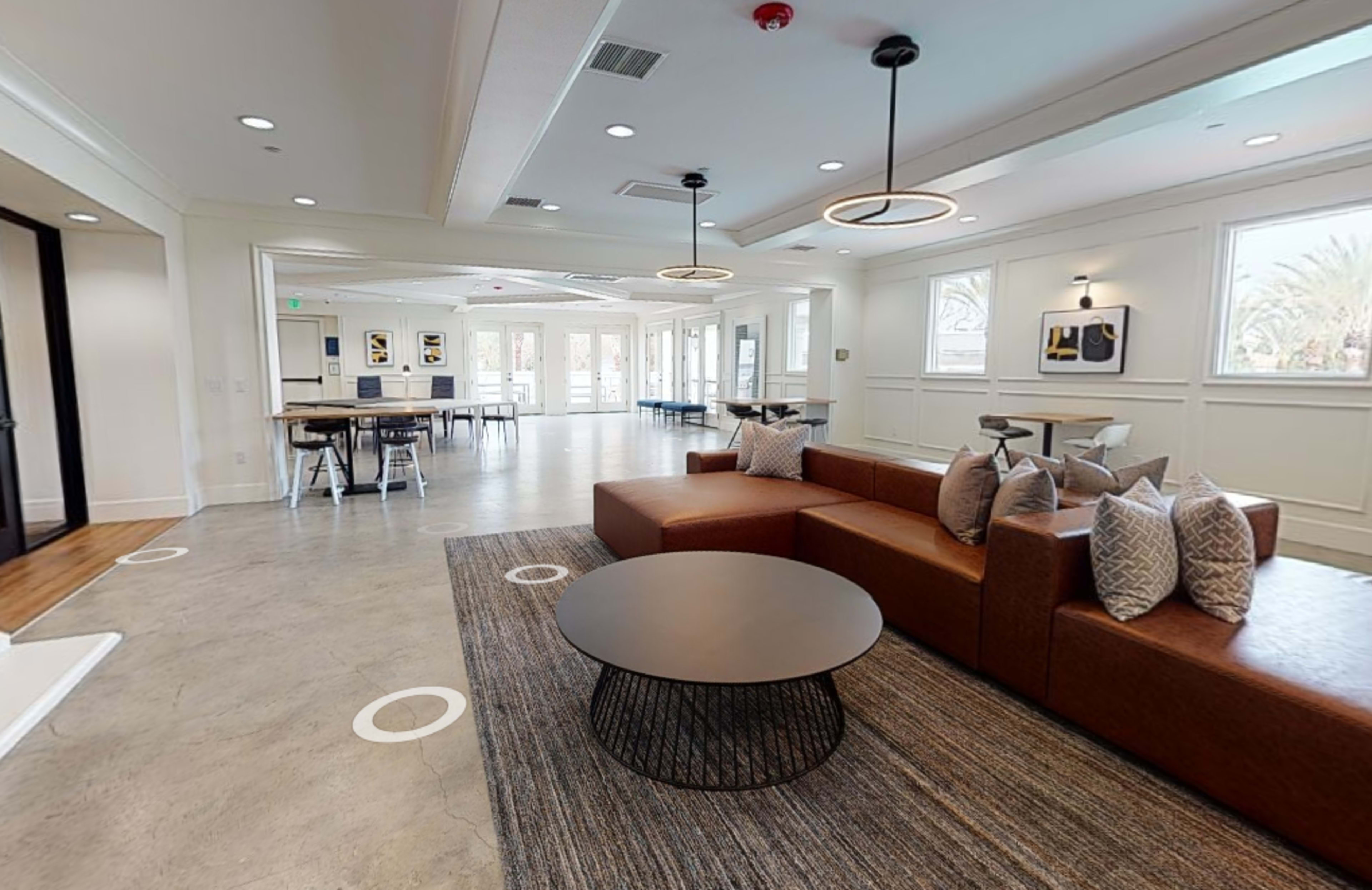 A modern lounge area featuring a brown sectional sofa, a circular coffee table, and tables with chairs arranged around a spacious, well-lit room with large windows.