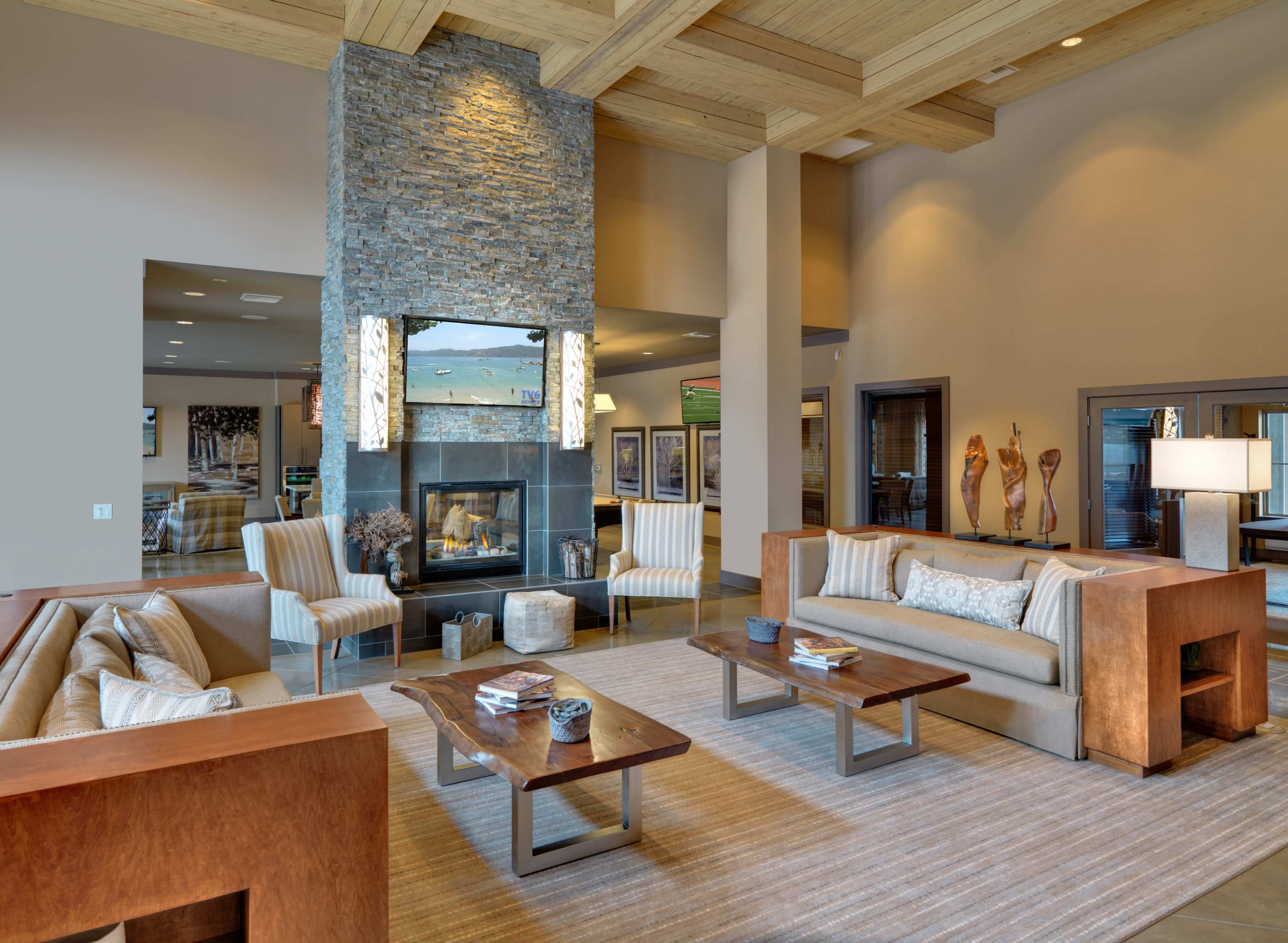 A spacious living room features two sofas, a central coffee table, and a stone fireplace with a mounted TV above it, surrounded by large windows and modern decor.