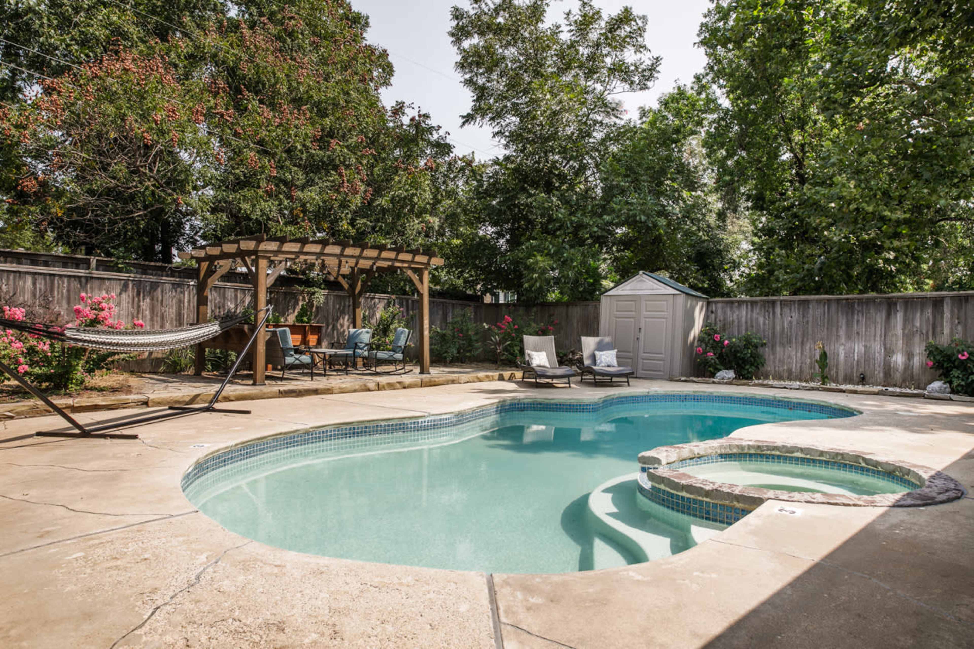 A landscaped backyard features a swimming pool with a built-in spa, surrounded by a wooden pergola, hammock, and seating area.