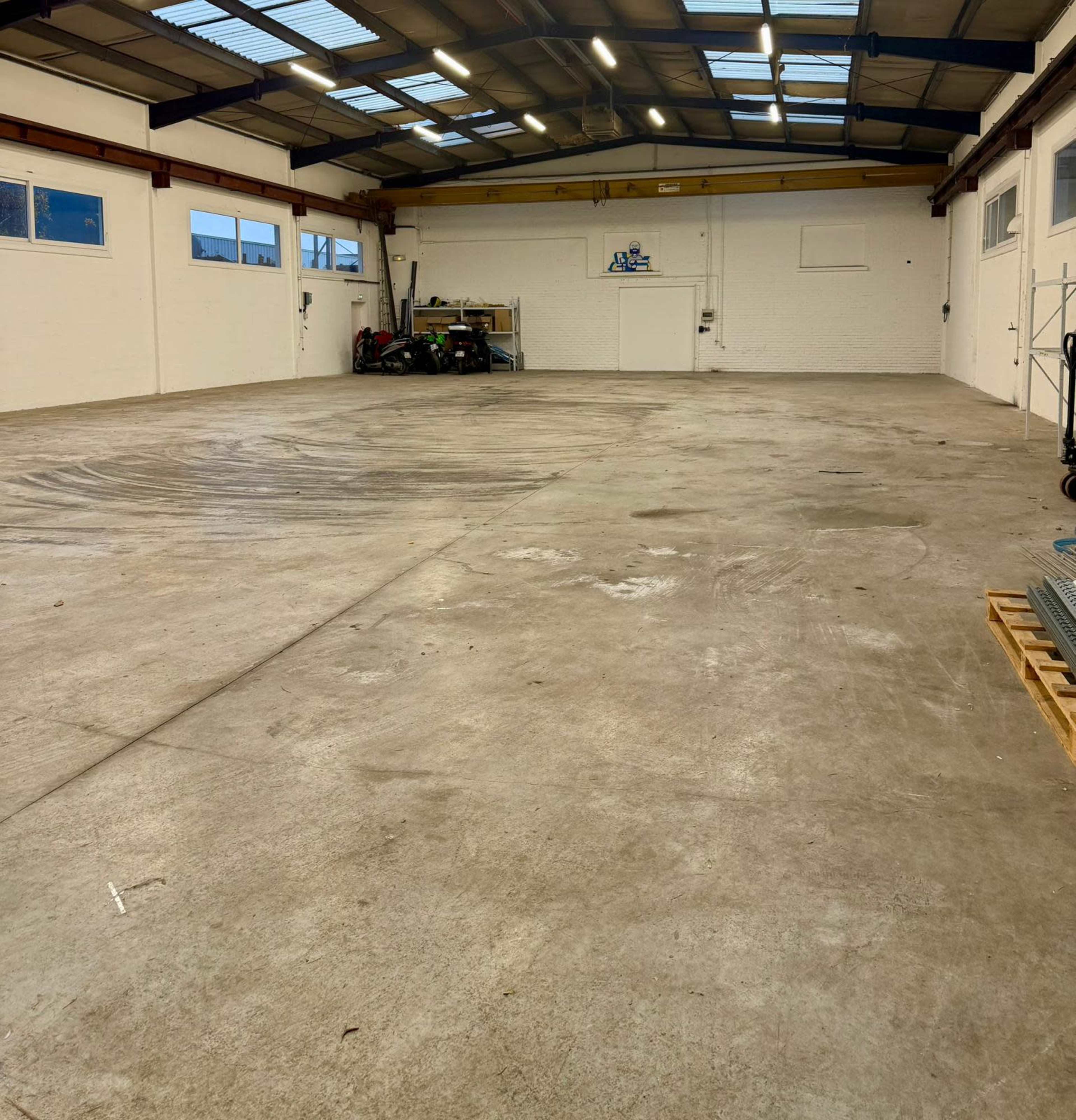 The image shows a large, empty industrial space with concrete flooring and high ceilings, featuring some equipment and storage against the walls.