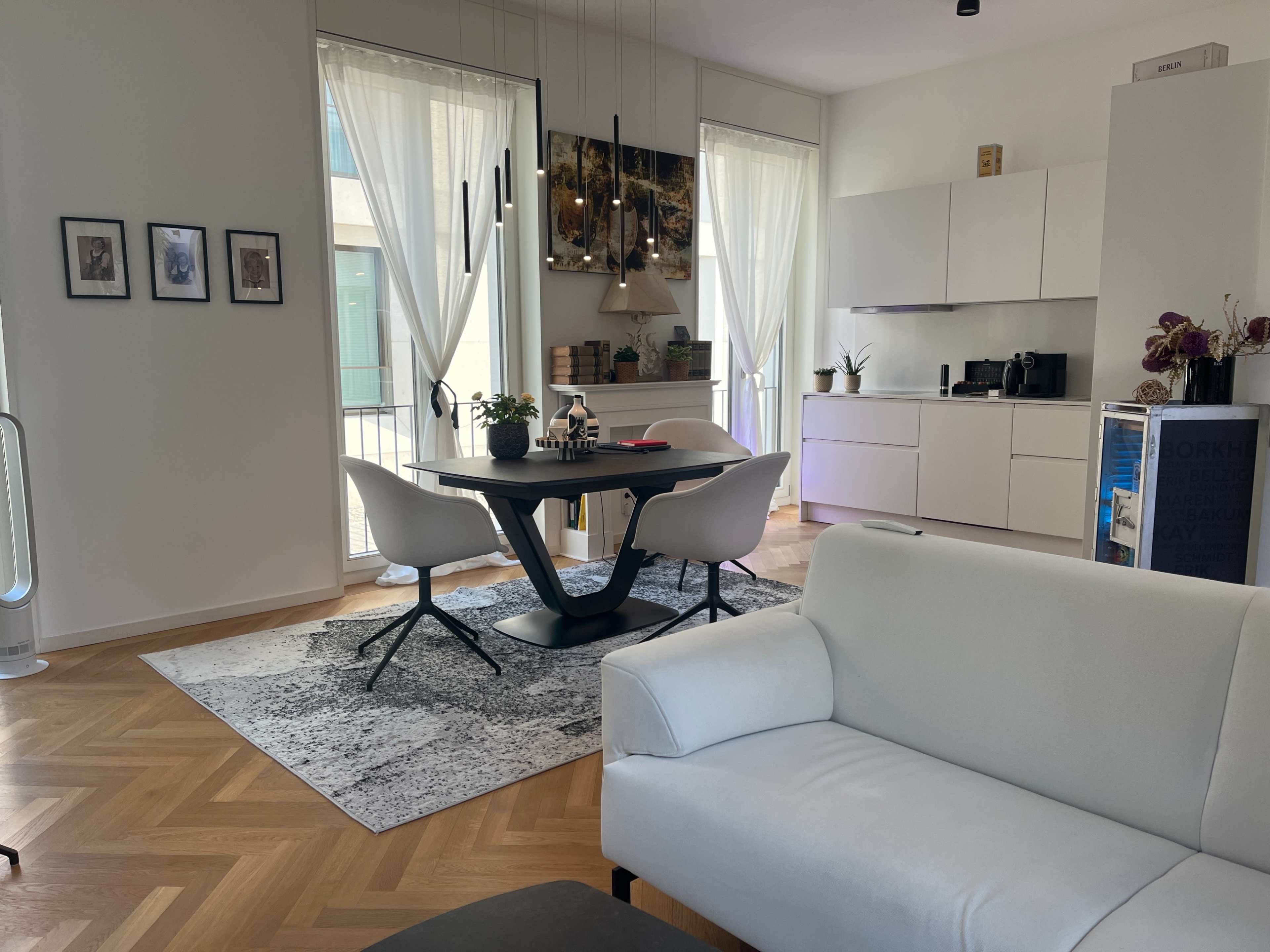 A modern living space featuring a dining area with a round black table and white chairs, a white sofa in the foreground, and a kitchenette with sleek cabinetry in the background.