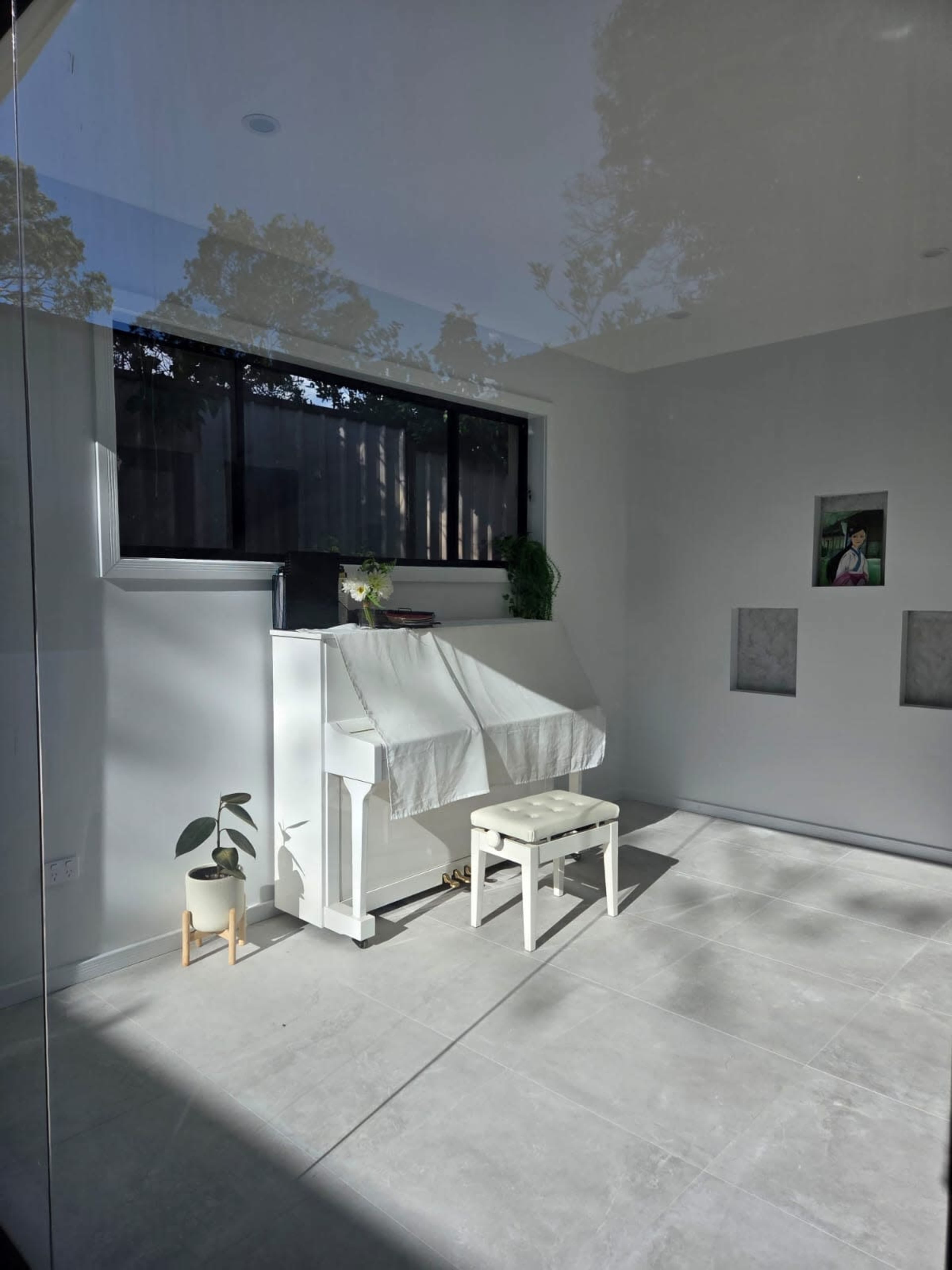A white piano and bench are positioned in a sunlit room with minimal decor and large windows.