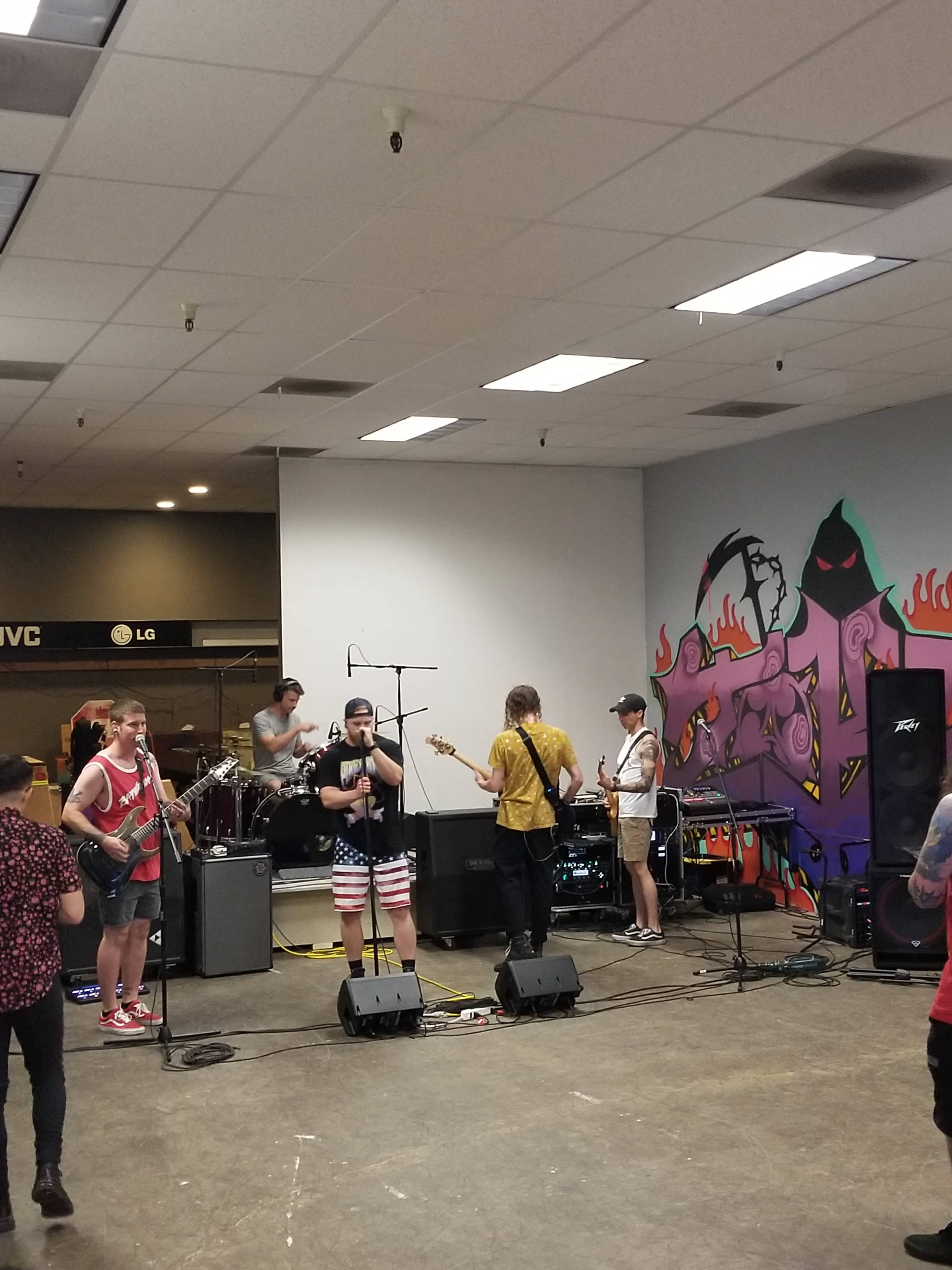 A band performs on a stage in a spacious room with colorful wall art and various musical instruments set up.