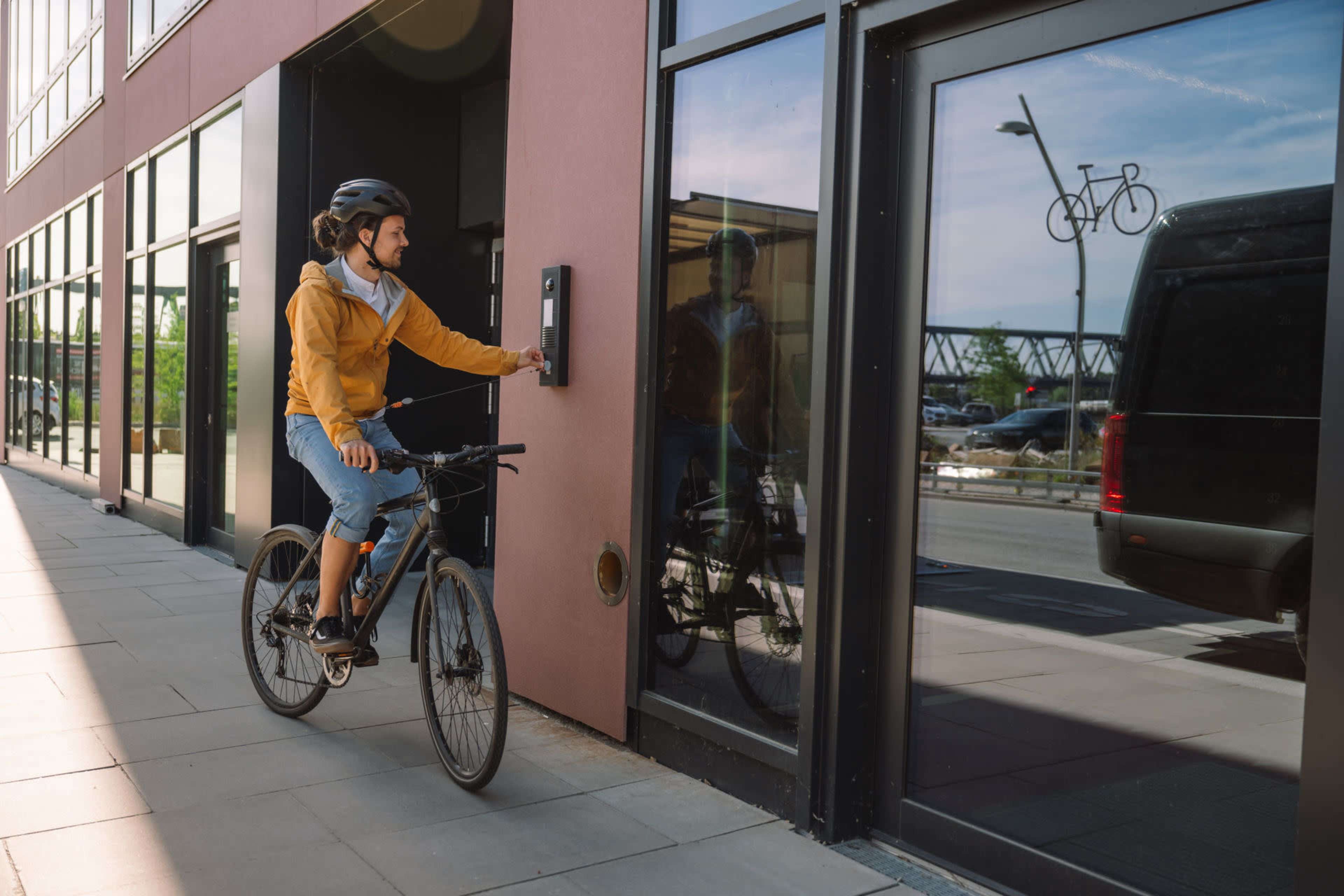 A person on a bicycle is pressing a call button next to a glass door of a building.