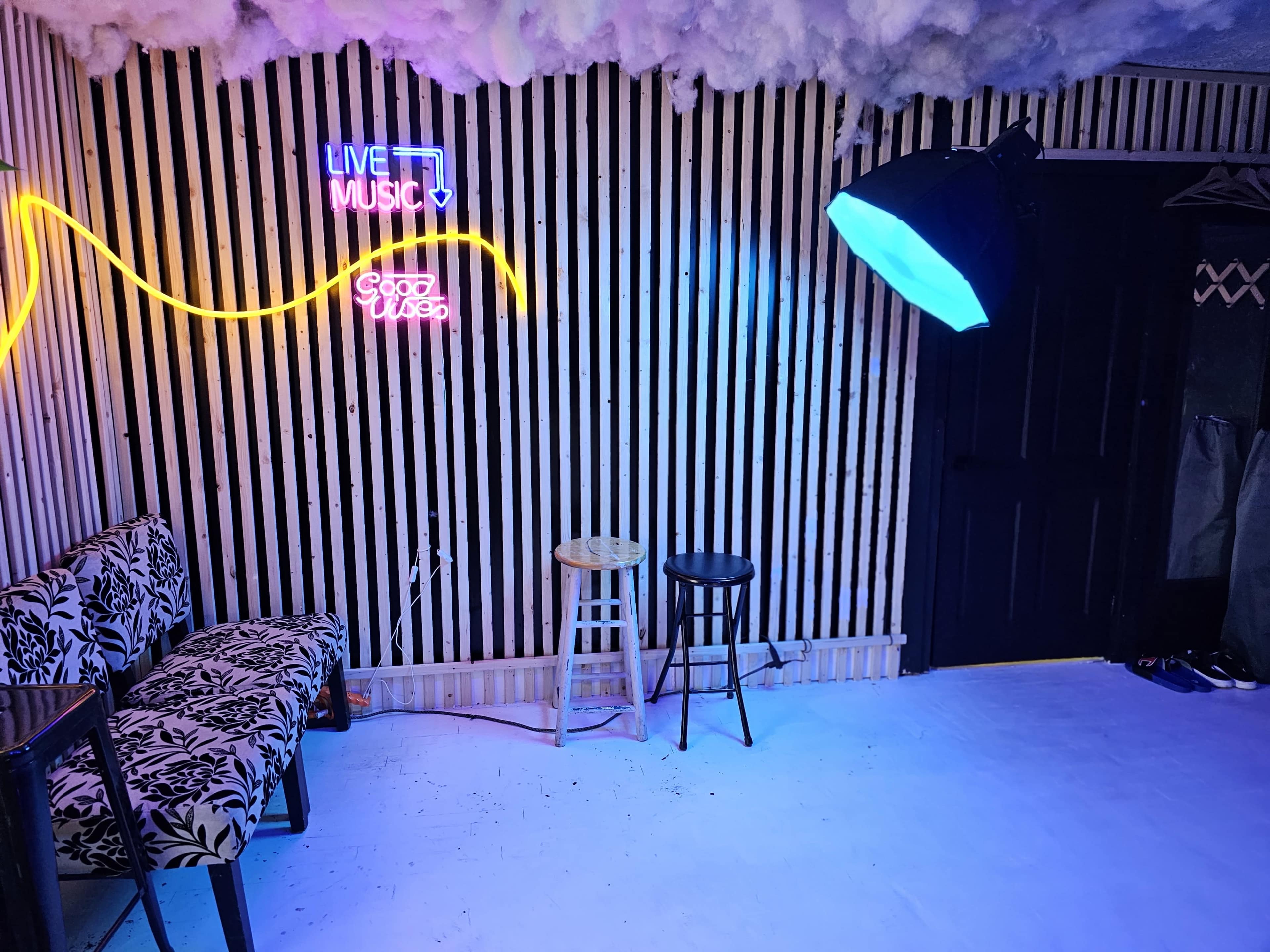 The image shows a small room featuring striped wooden walls, a neon sign that reads "LIVE MUSIC," a chair and a stool, and a large overhead light.