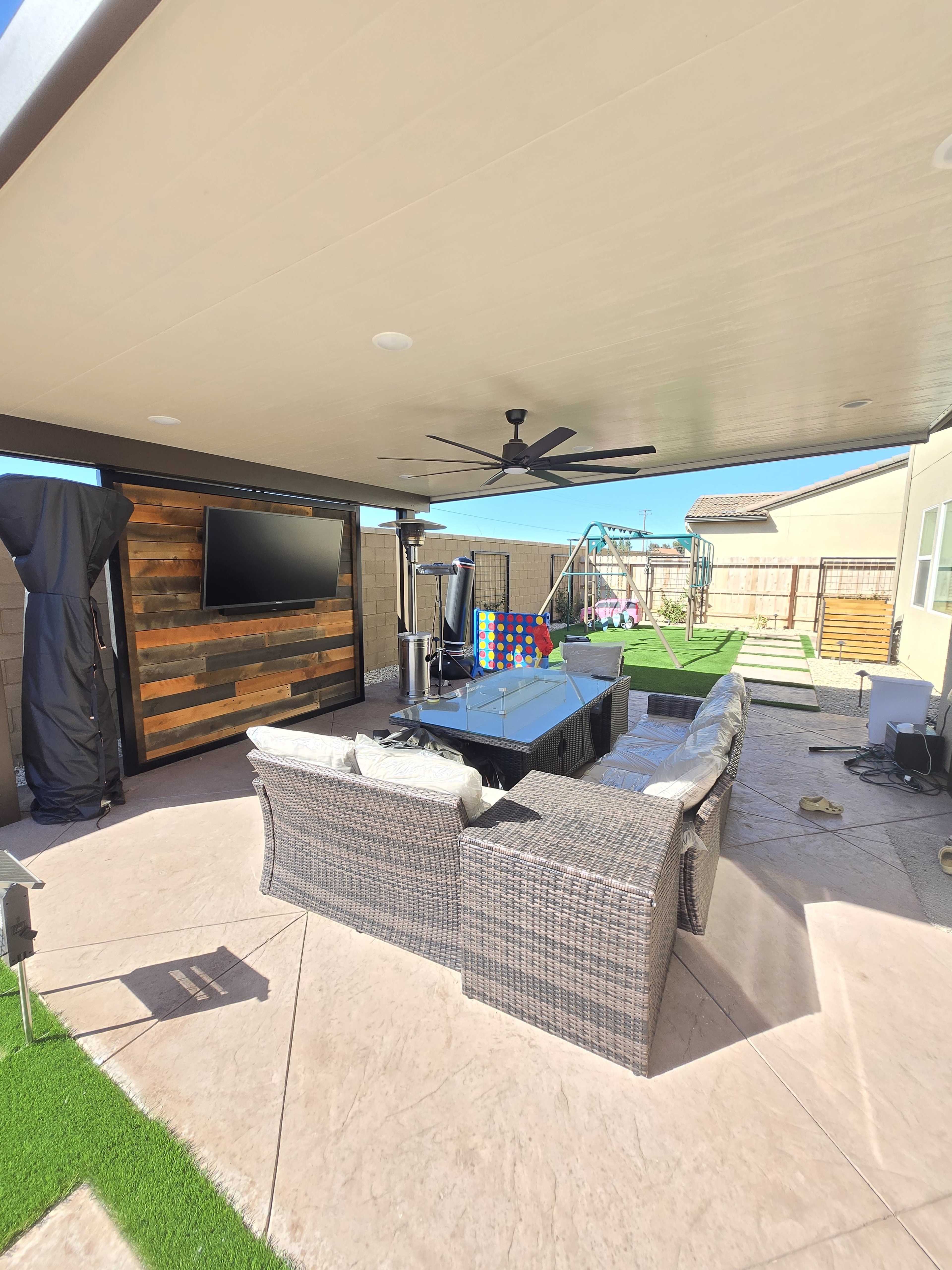 The image shows a covered outdoor patio with a sectional sofa, a television mounted on a wooden wall, and a playground area in the background.