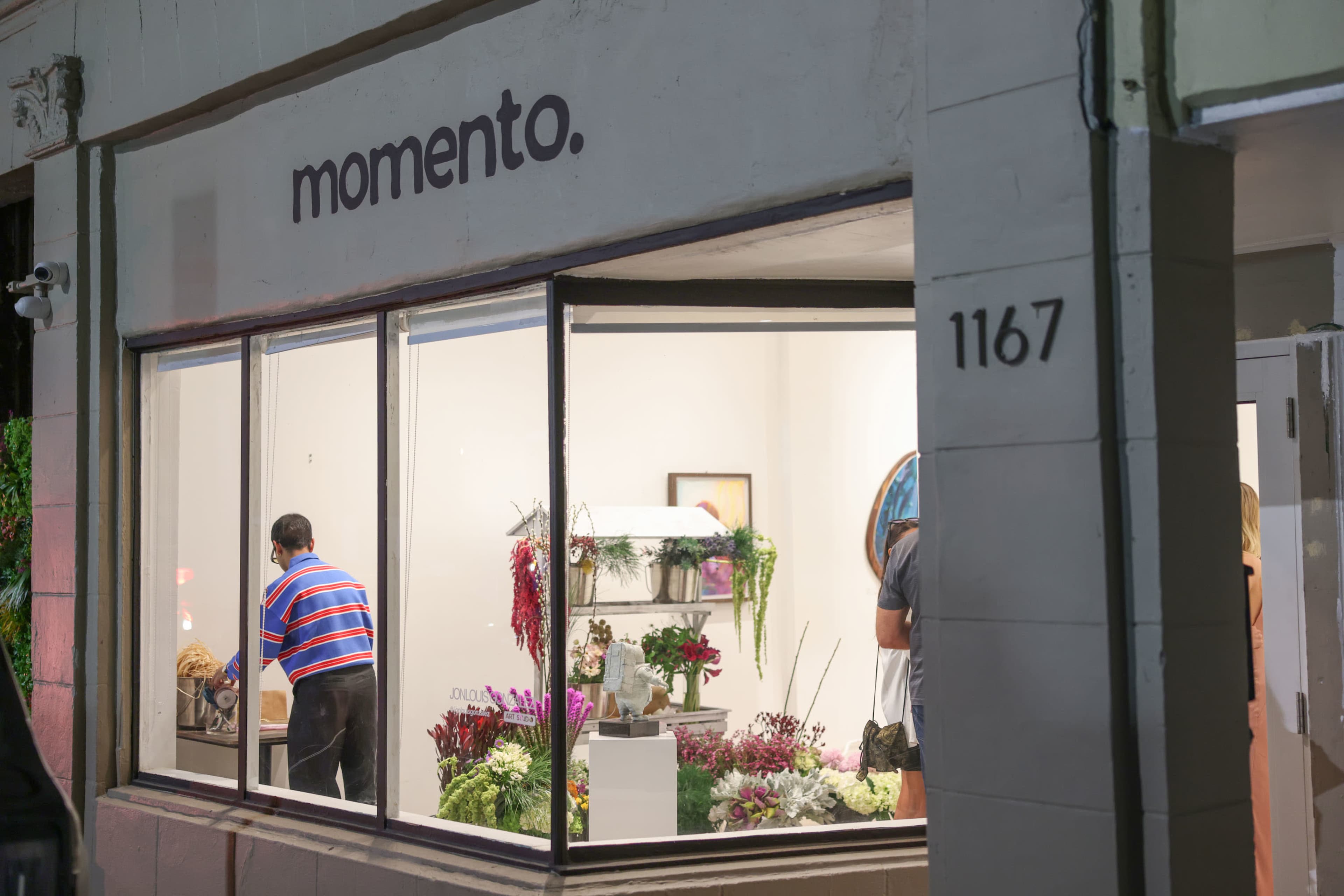 The image shows a storefront named "momento" with a large window displaying floral arrangements and two people inside, one arranging flowers and another standing in the background.