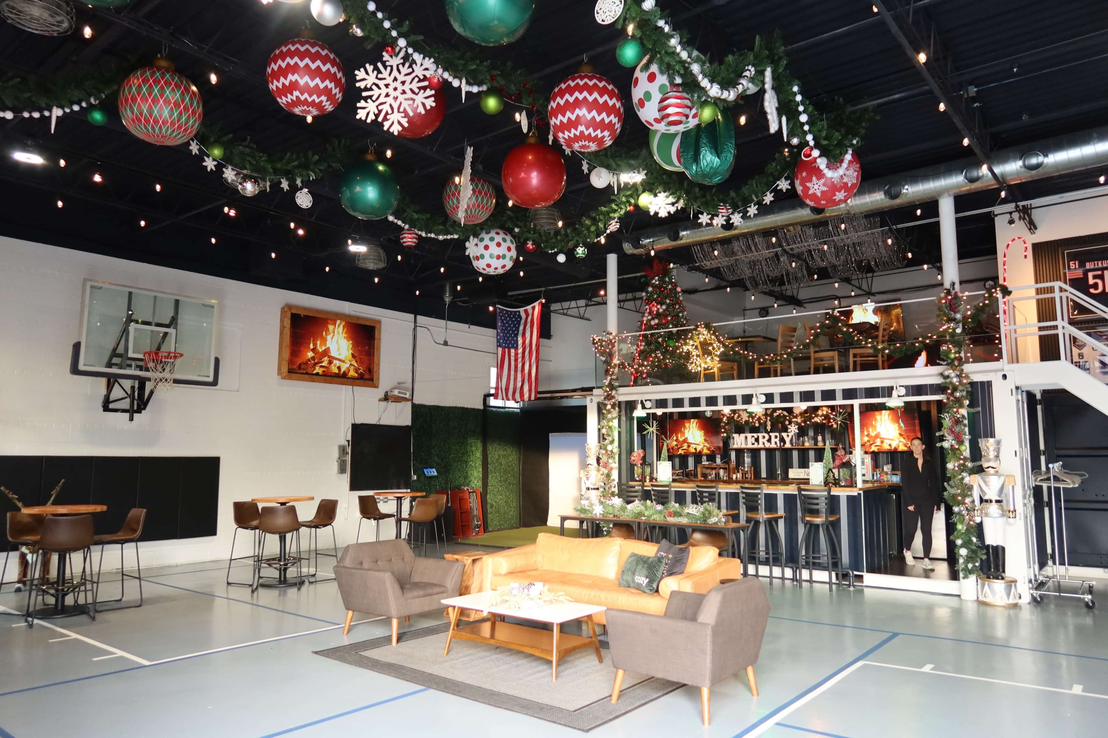 The image depicts a large, festive indoor space featuring Christmas decorations, including ornaments hanging from the ceiling, a decorated tree, and a cozy seating area with a basketball hoop on the wall.