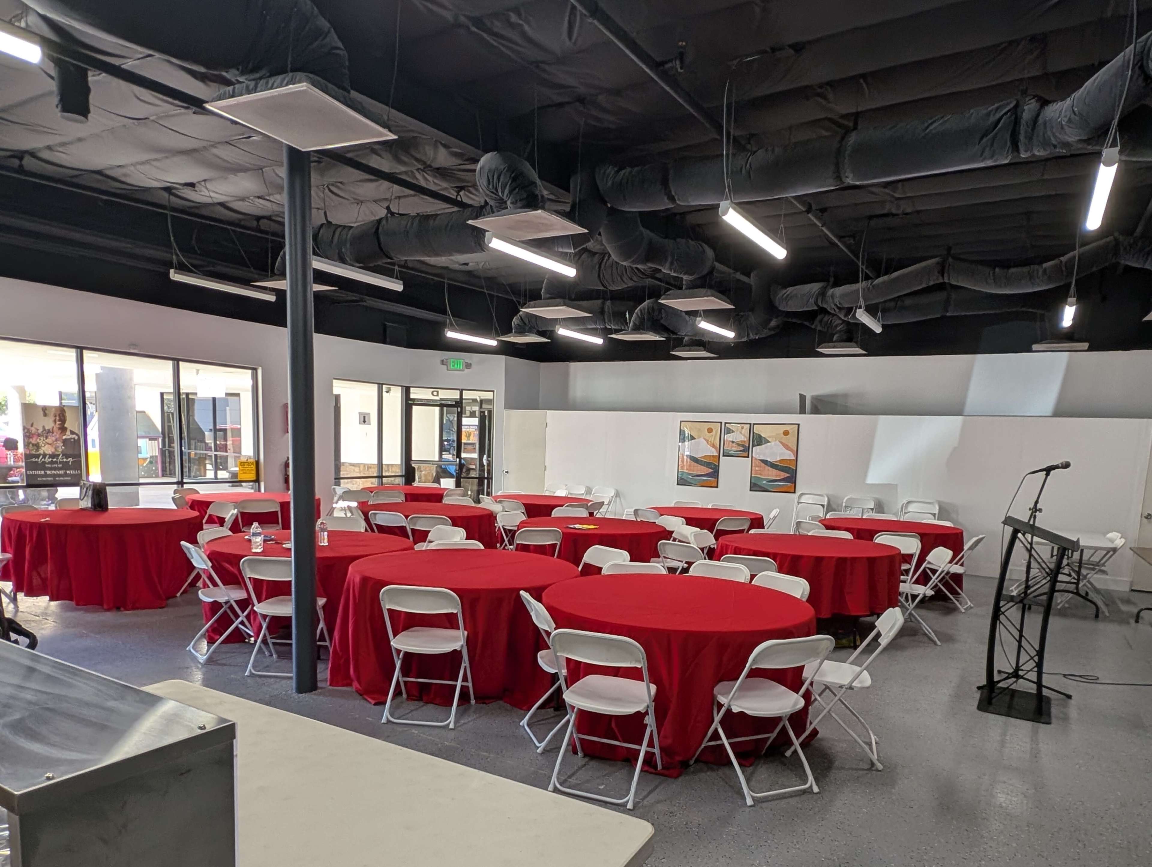 The image shows a spacious event room arranged with several round tables covered in red tablecloths and white chairs, alongside a podium at the front.