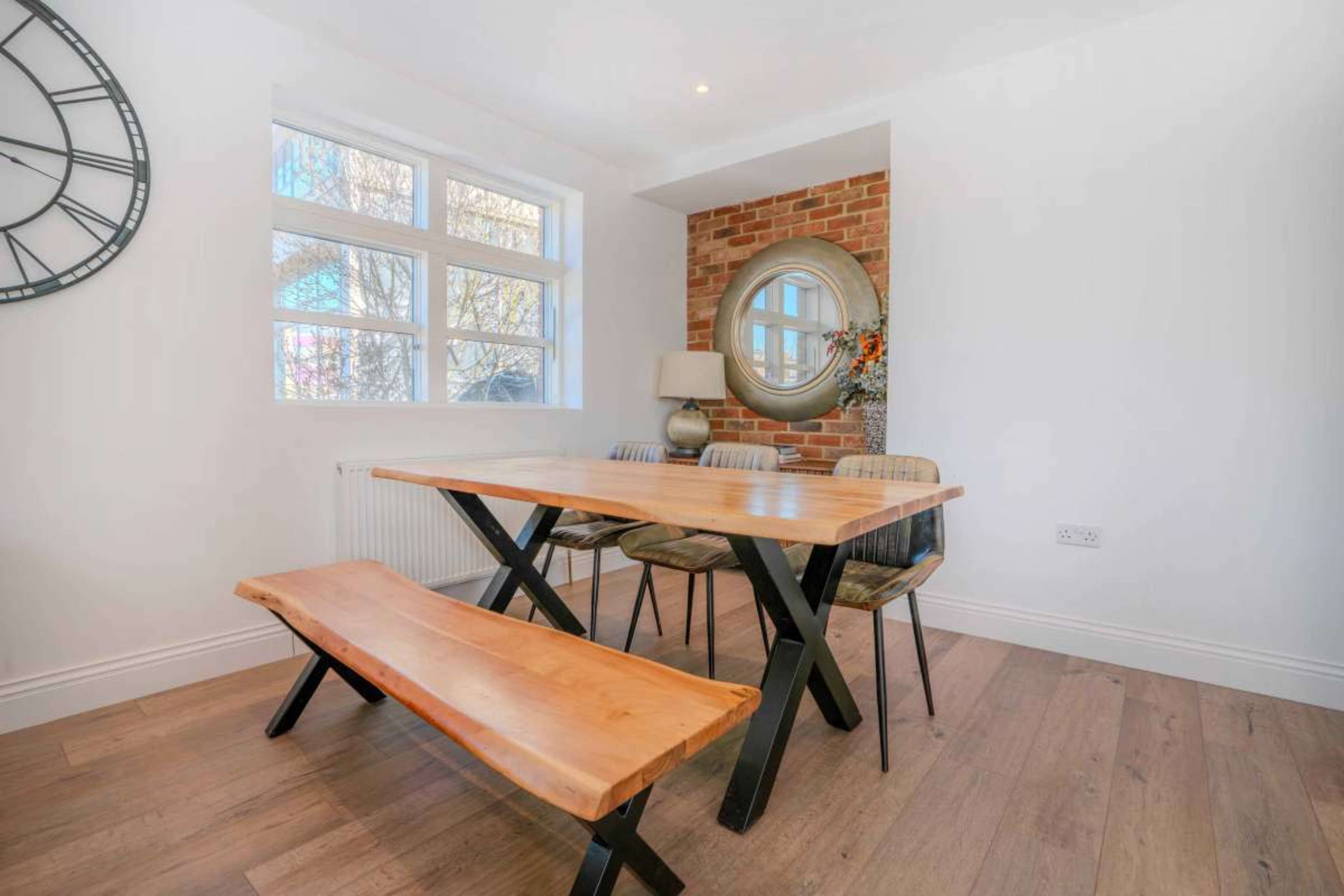 A wooden dining table with a matching bench and chairs is positioned in a bright room featuring a brick wall and a round mirror.