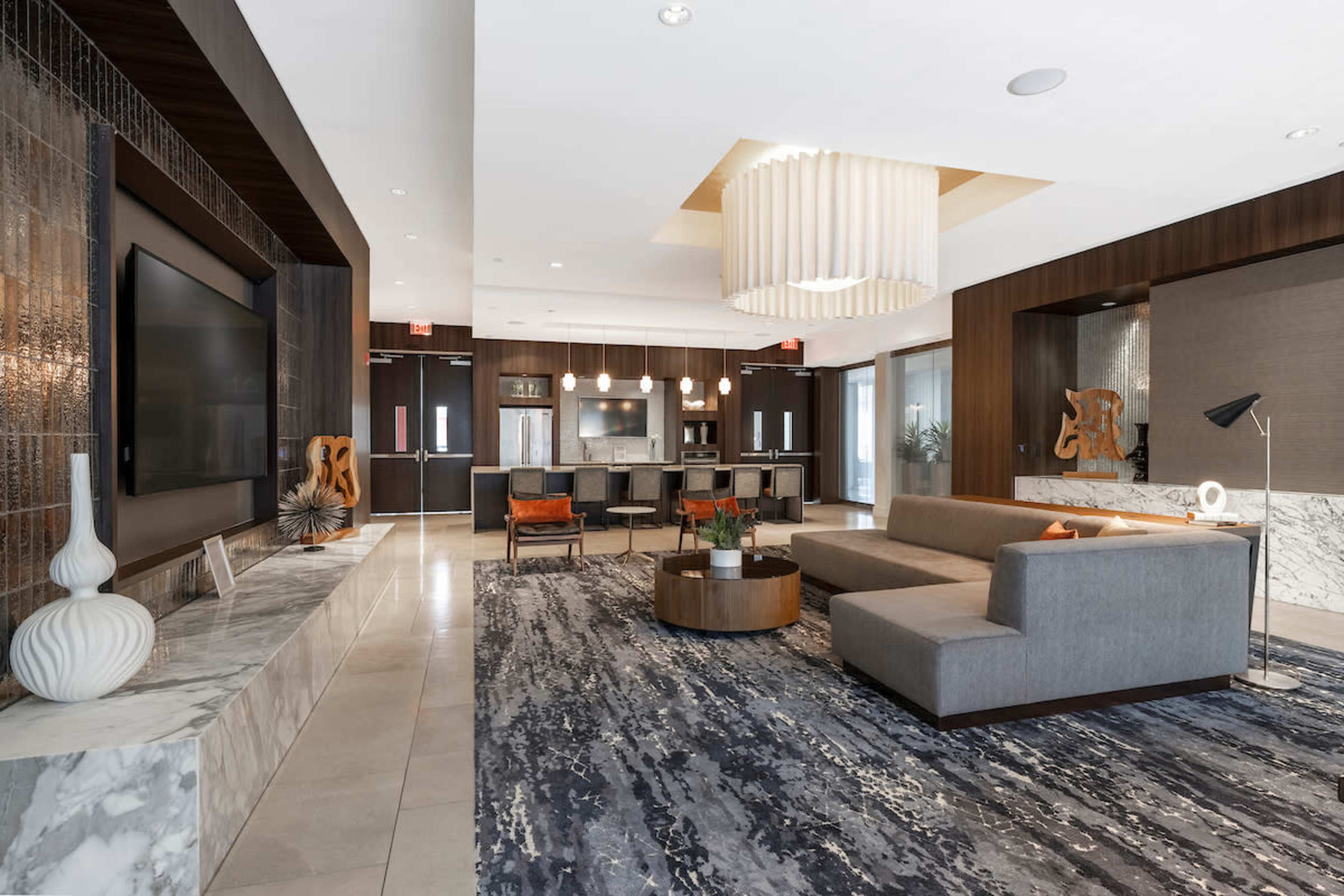 A modern lobby features a gray sofa, marble surfaces, a large television, and a bar area with stools.
