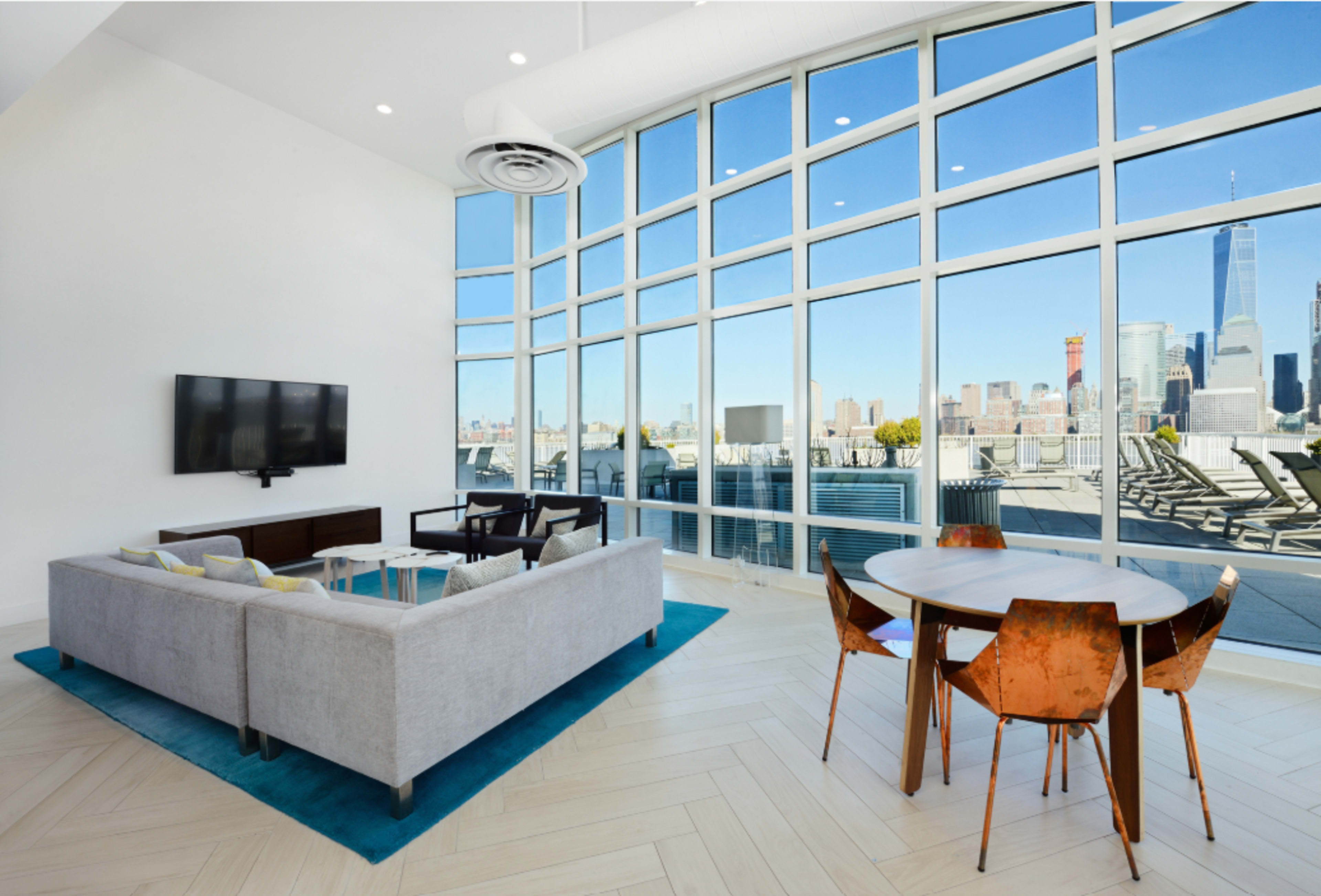 The spacious room features a modern living area with a gray sofa, a glass table, and large windows showcasing a city skyline.