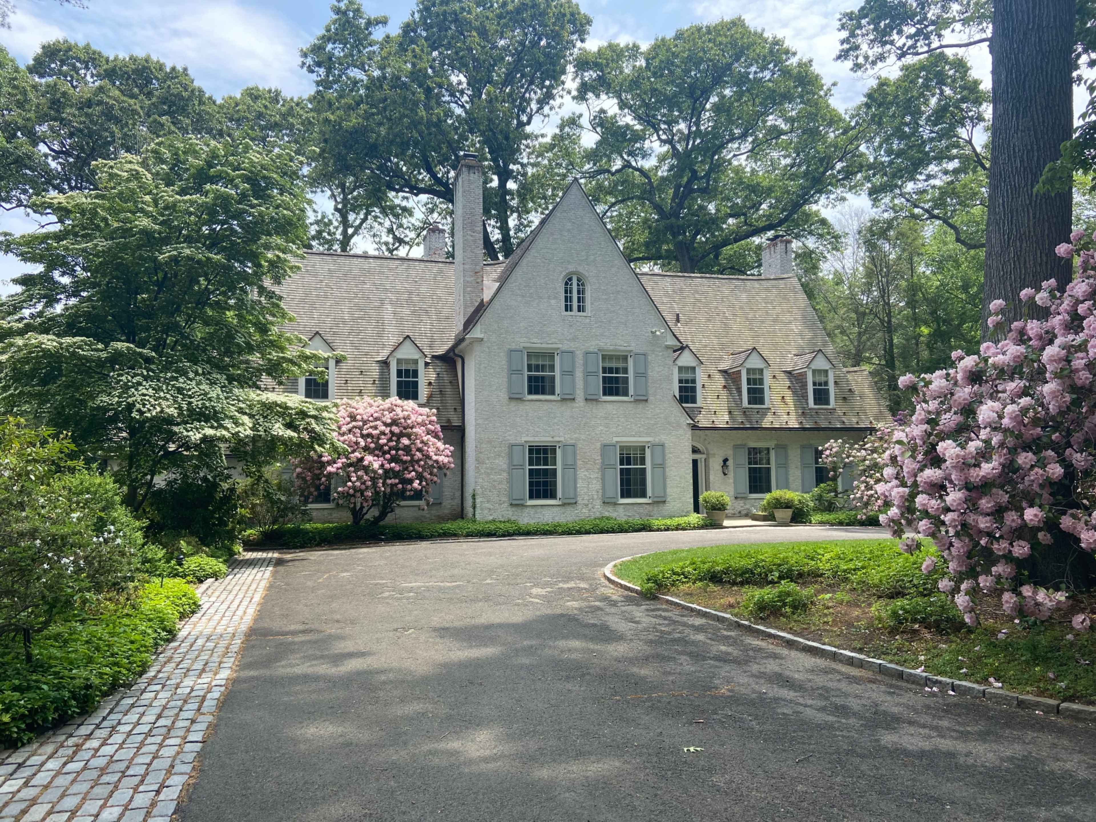 The image shows a large, spacious white house with a steeply pitched roof, surrounded by greenery and flowering bushes along a paved driveway.