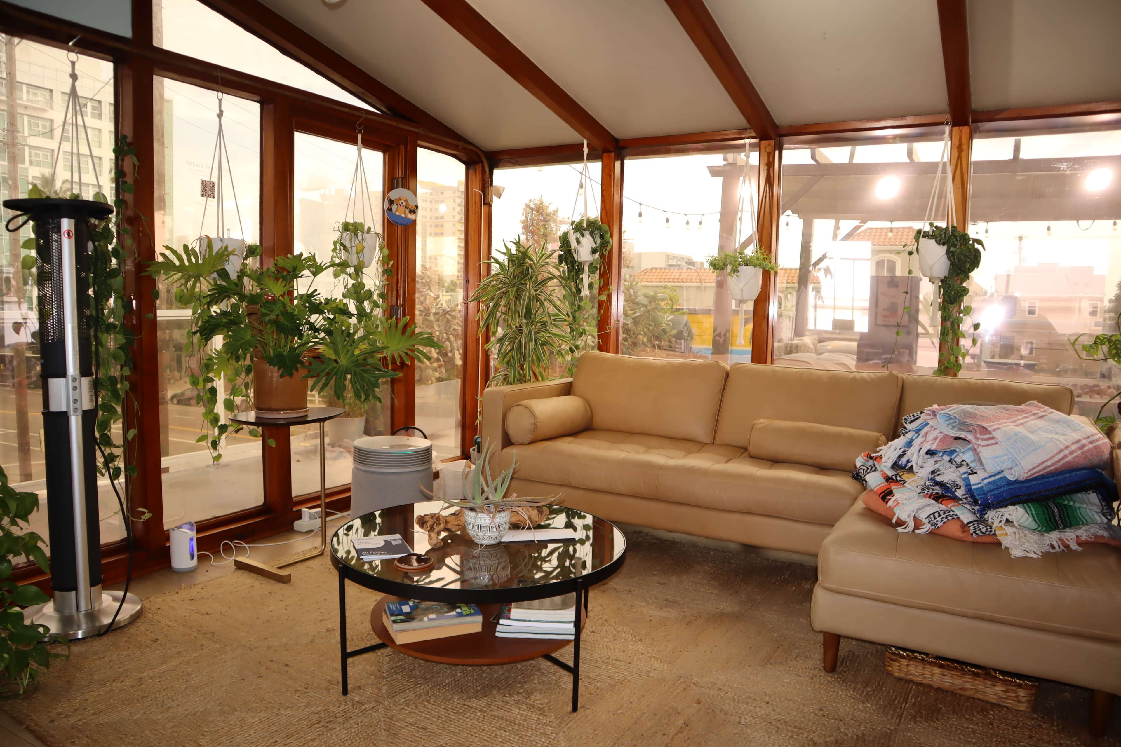 A sunlit room features a beige couch, a round glass coffee table, potted plants, and a stacked collection of blankets.