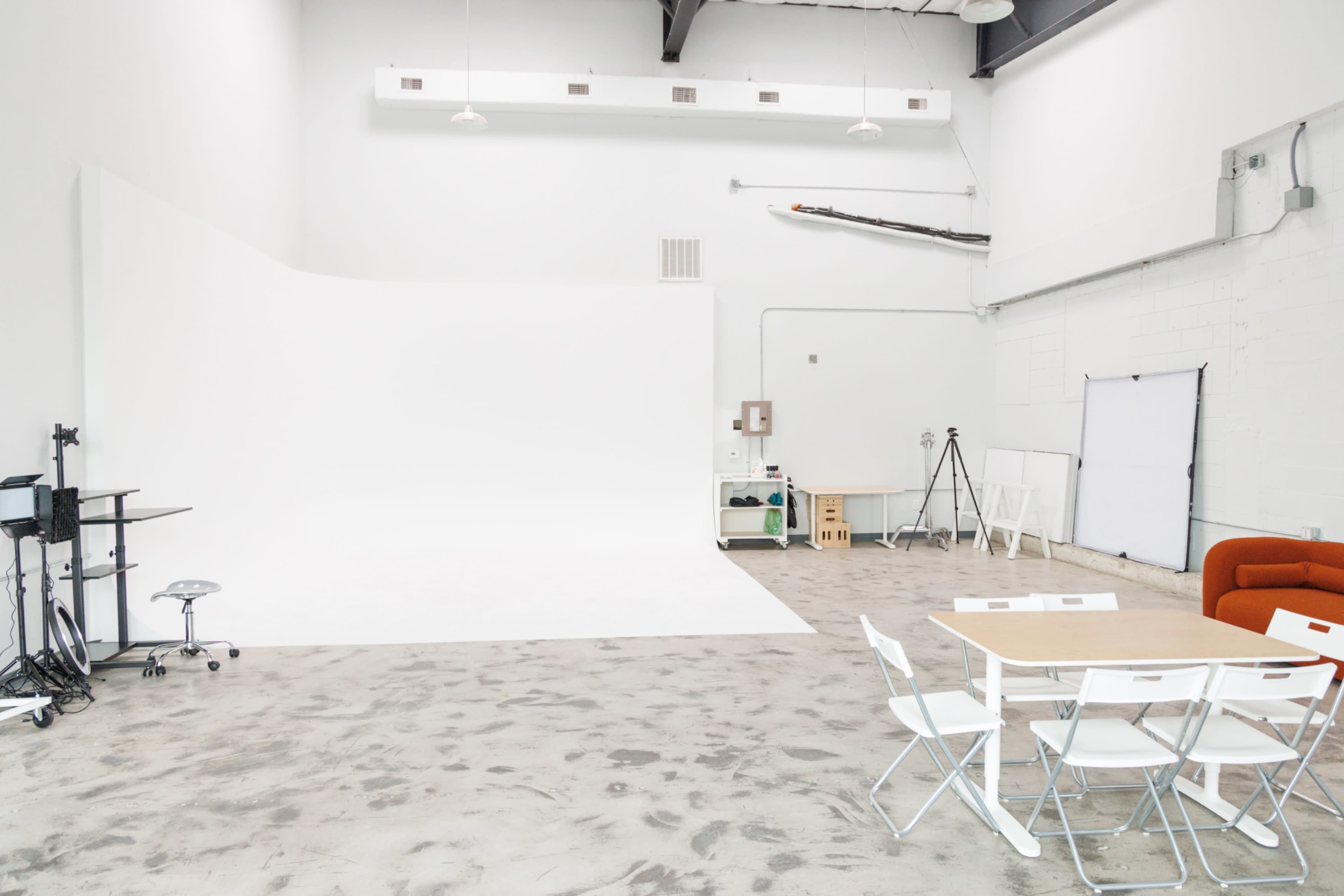 The image shows a spacious white studio with a clean backdrop, a table with chairs, and various equipment arranged in the corners.