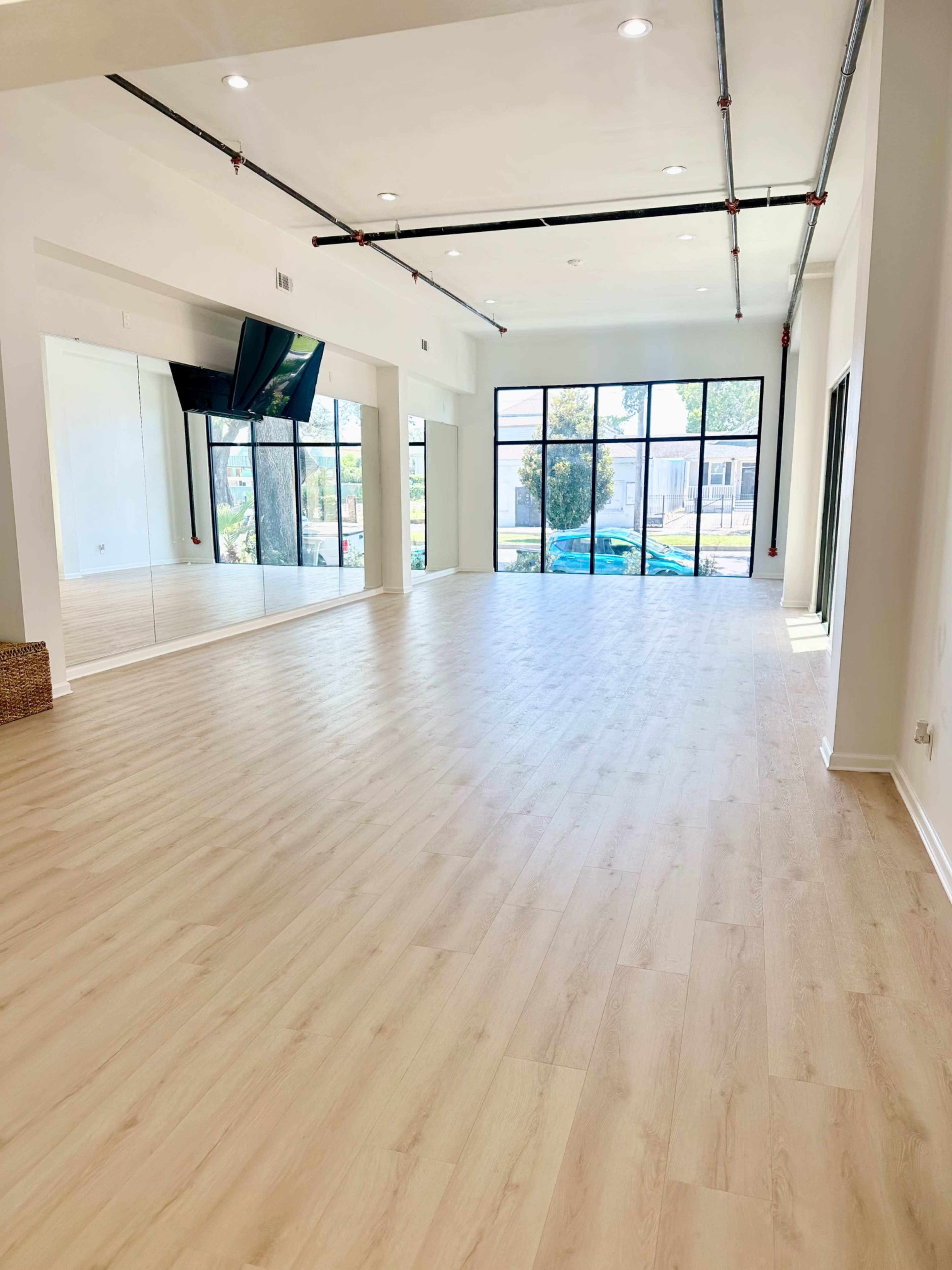 The image shows a spacious, empty room with large windows and a wooden floor, featuring a mirrored wall on one side.