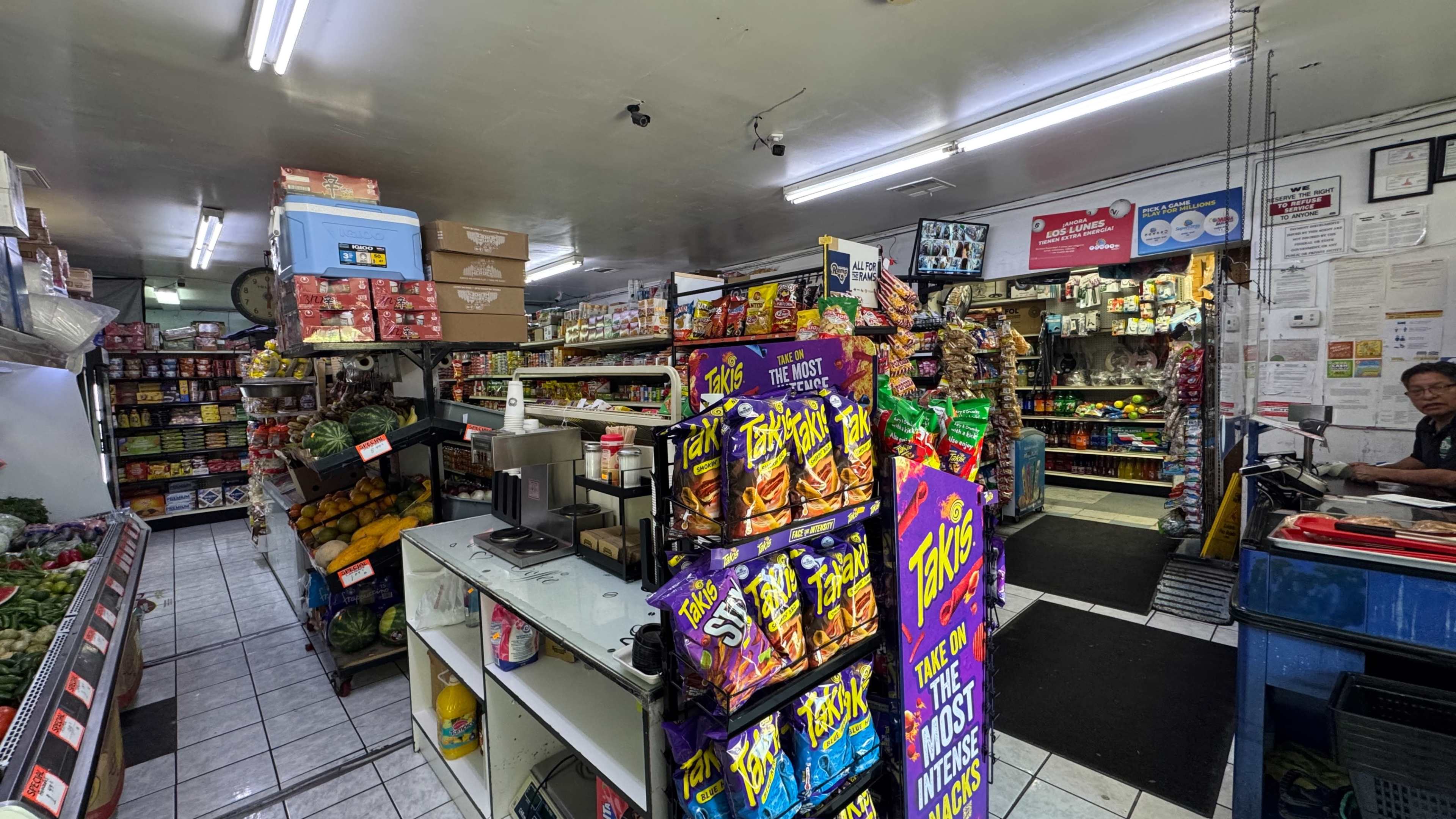 The image shows an interior of a small grocery store filled with various food items, snacks, and produce displayed on shelves and in baskets.