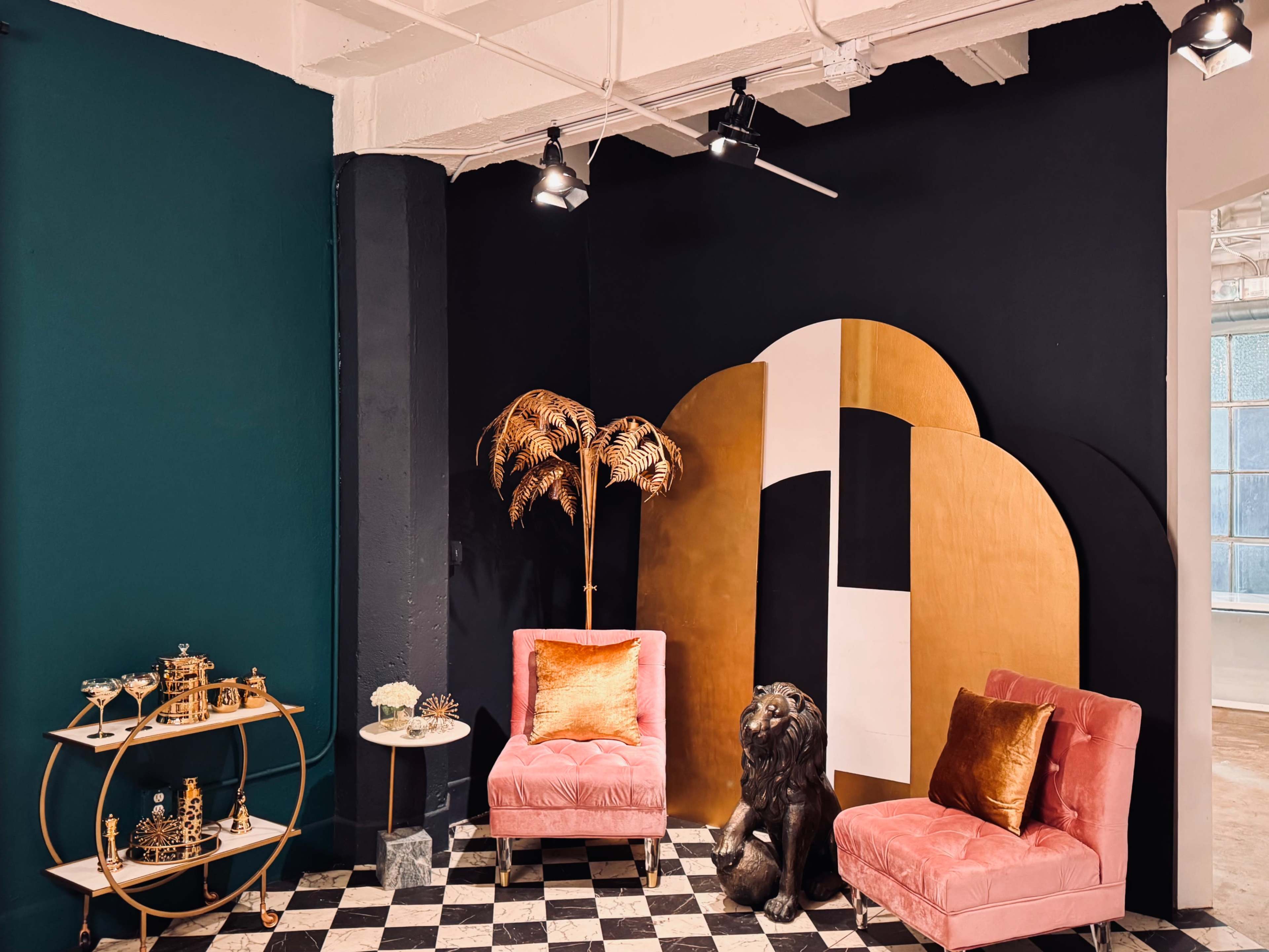 The image shows a stylish corner of a room featuring two pink armchairs, a decorative gold and white arch, a black and white checkered floor, and a small table with a cocktail cart and a floral centerpiece.