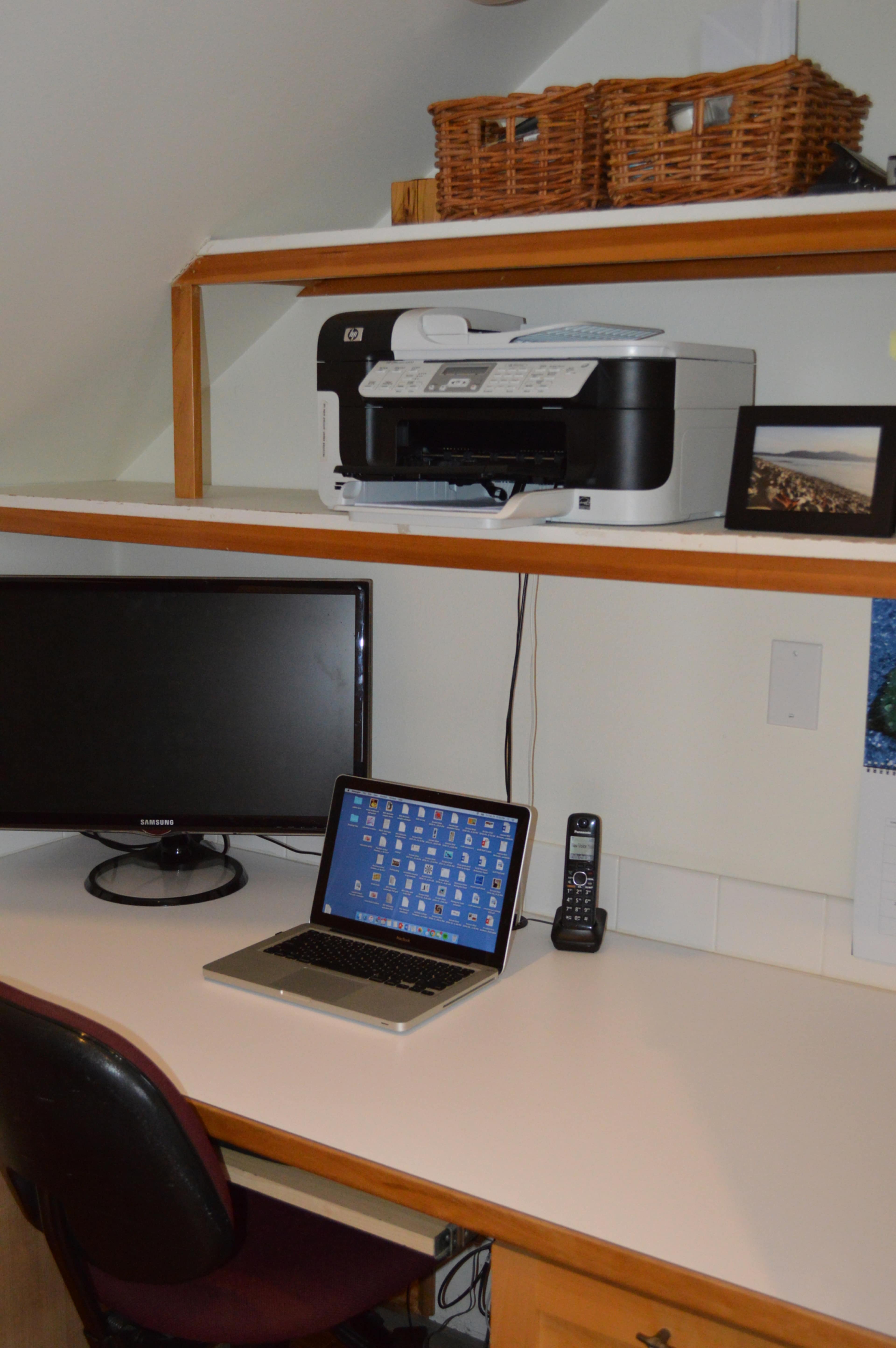 The image shows a home office setup featuring a printer on a shelf, a computer monitor and a laptop on a desk, along with a telephone and a framed photo.