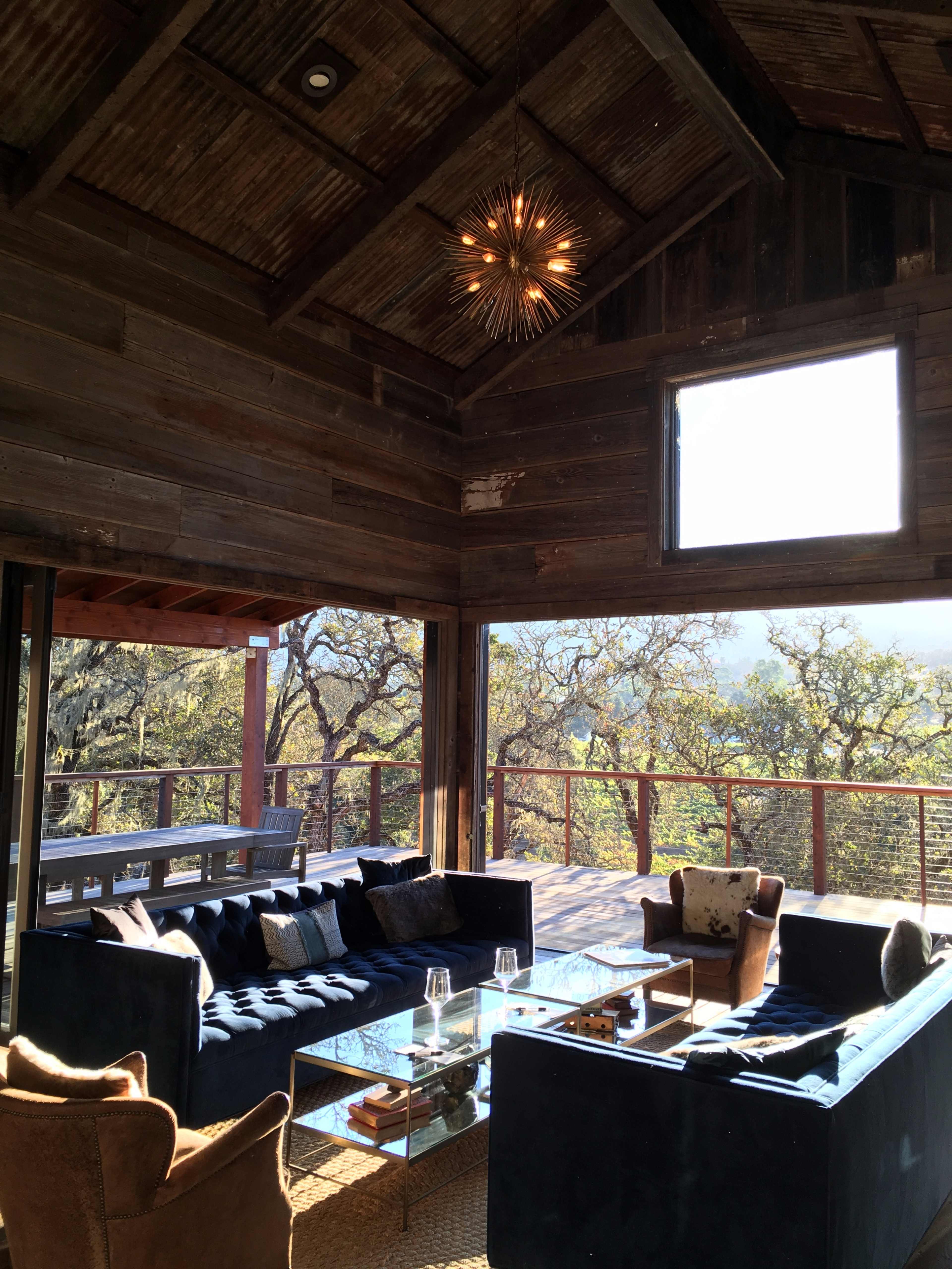 A cozy living area with large windows, featuring dark sofas, a glass coffee table, and a decorative light fixture, all set against a backdrop of trees visible outside.