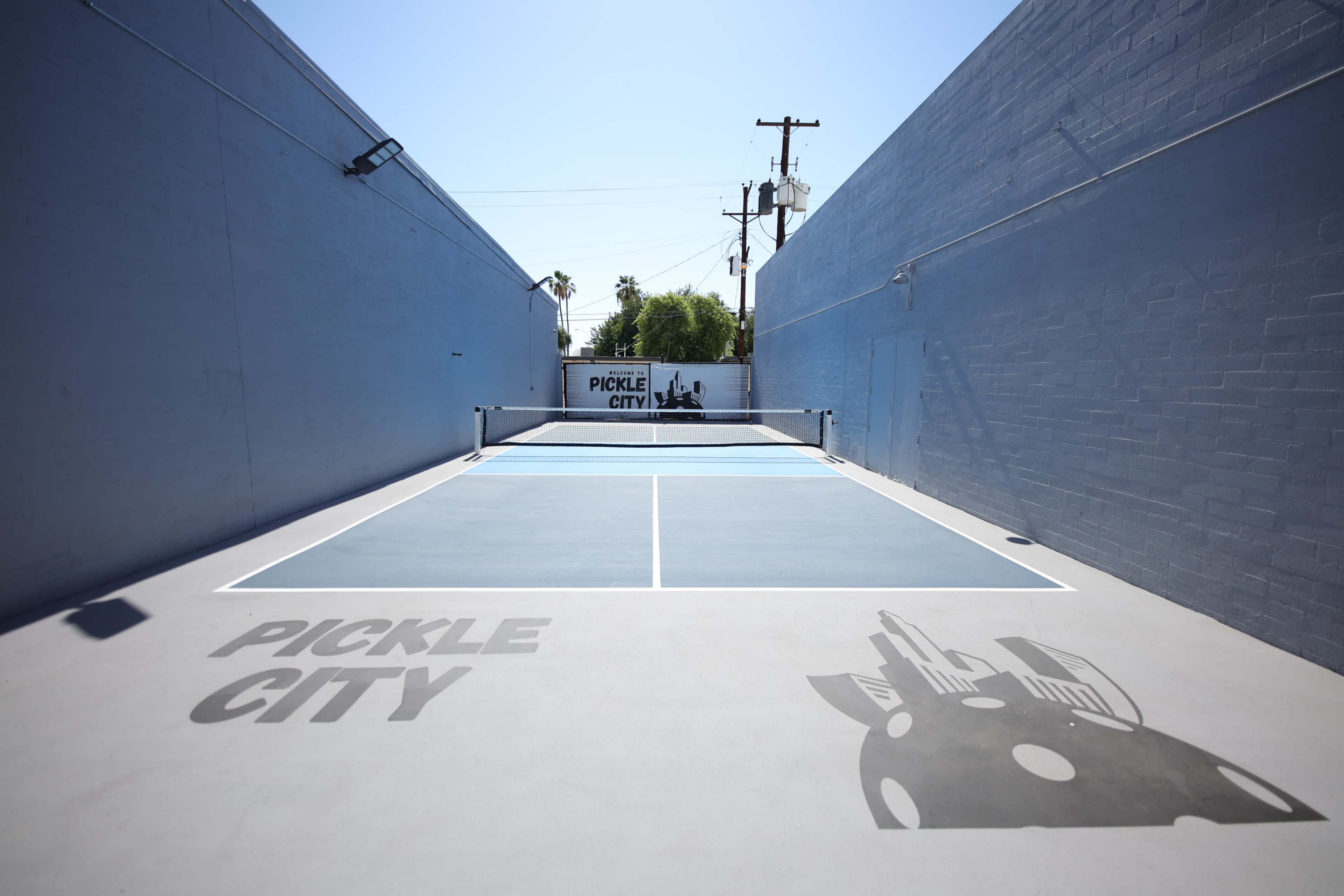 A pickleball court with clear blue surfaces is situated between two gray walls, featuring the words "PICKLE CITY" displayed prominently.