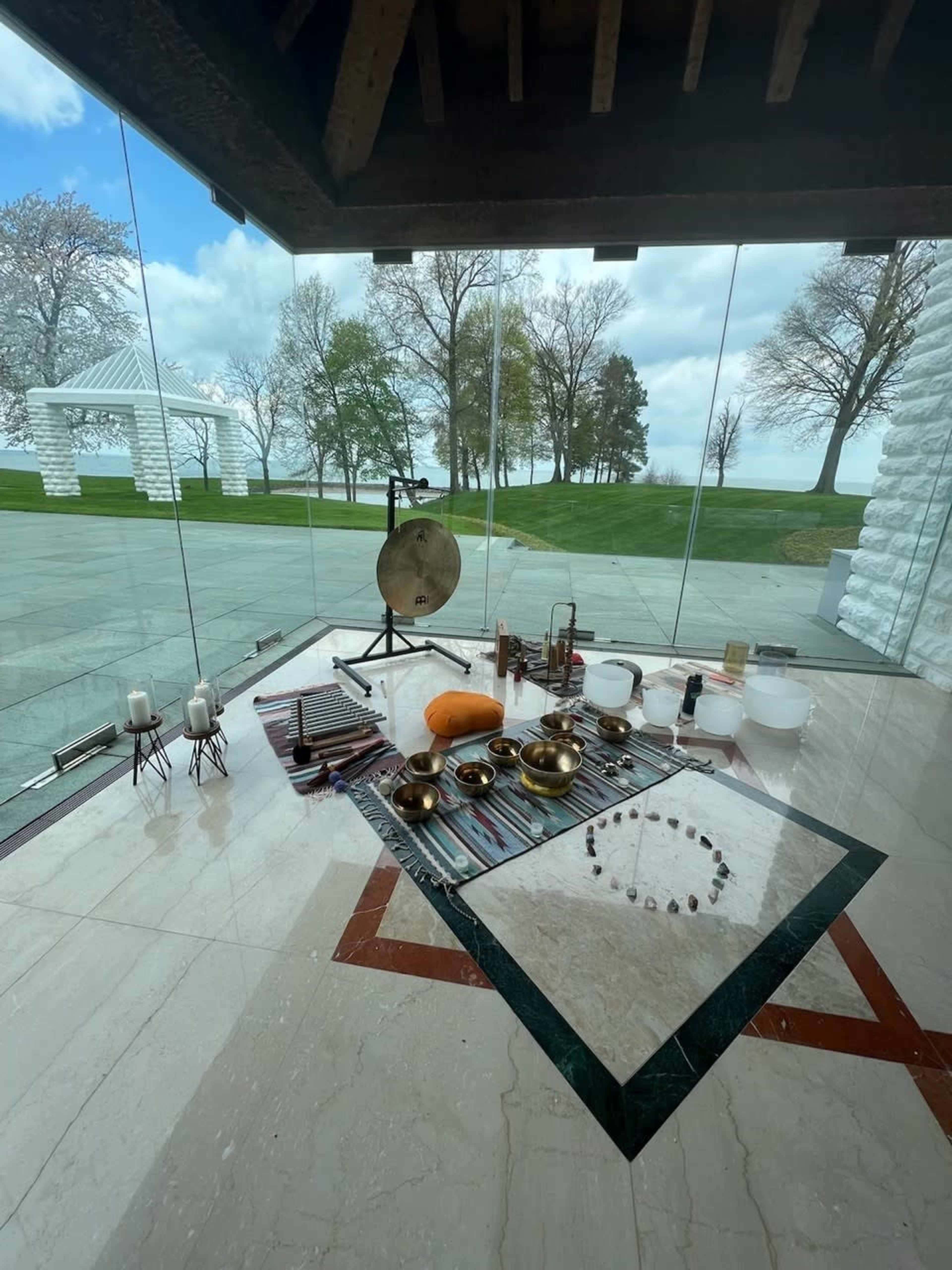 The image shows a reflective glass room featuring various musical instruments arranged on a decorative mat, alongside candles and a large gong, with a view of a landscaped area outside.