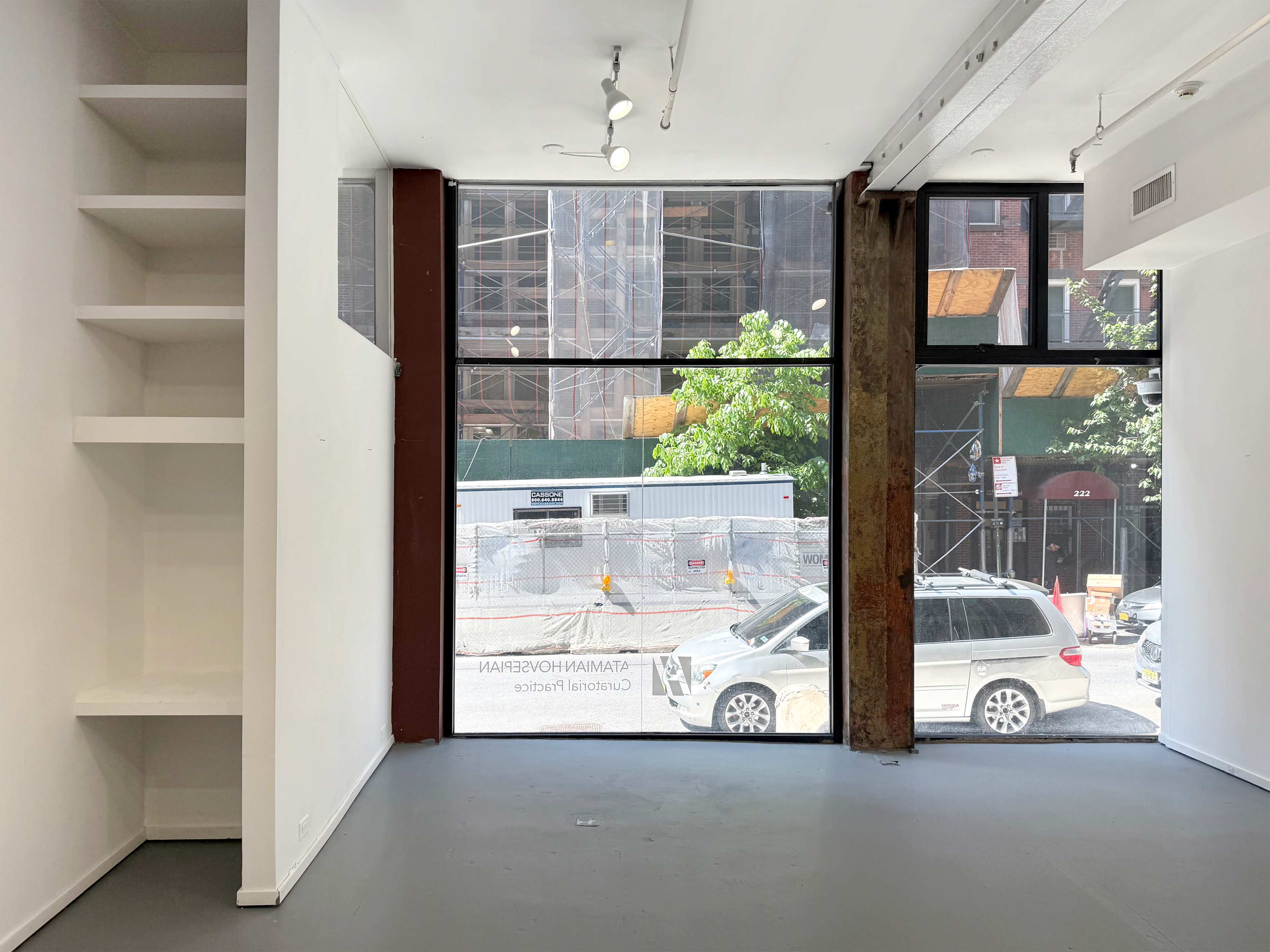 The image shows an empty interior space with a large window facing a construction site, featuring white walls and a built-in shelf on the left.