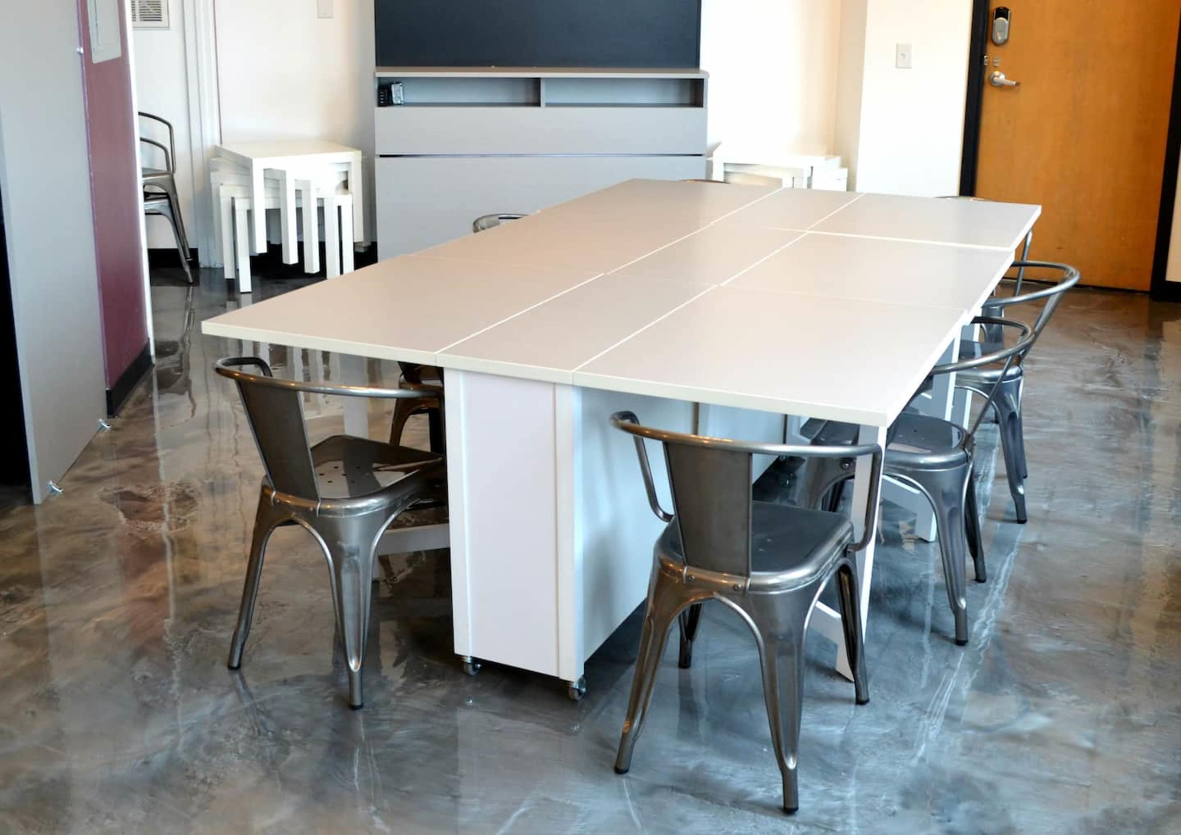 A modern conference room with a large white table surrounded by several metallic chairs.